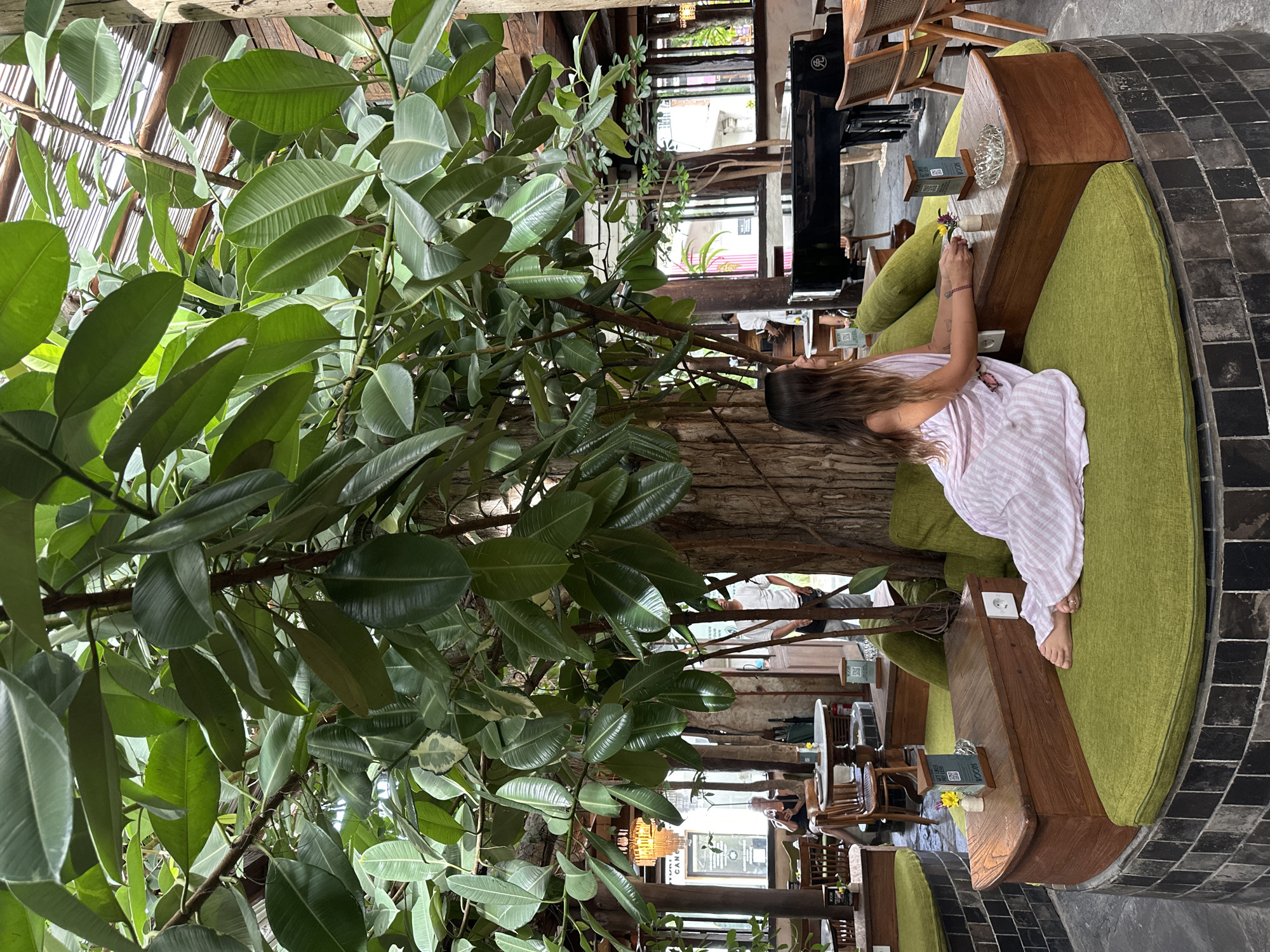 This media file depicts a tranquil cafe or restaurant setting in Mengwi, Indonesia, characterized by its integration of natural elements. The main subject is a woman with long, dark hair, wearing a light pink and white striped dress, seated barefoot on a lime-green upholstered sofa. She is positioned next to a large, leafy tree with a thick trunk and broad green leaves that stands prominently within the establishment, creating a lush, organic atmosphere.
The environment combines rustic wooden tables and seating with dark, irregularly shaped stone floor tiles. The architecture features exposed wooden beams and slatted structures overhead, suggesting a semi-open-air design that allows natural light to filter through, indicating it is daytime. In the background, other patrons are visible, seated at various tables, and large window-like openings offer glimpses of the outside.
The woman appears to be relaxing, with her hands near a small vase holding a flower on the wooden table next to her. An ashtray and a small card are also on the table. A menu or sign is visible with a "SCAN ME" QR code, and partially legible text that includes "CAN" and "FOODS." The scene conveys a sense of peace and comfort, typical of a tropical and natural retreat.