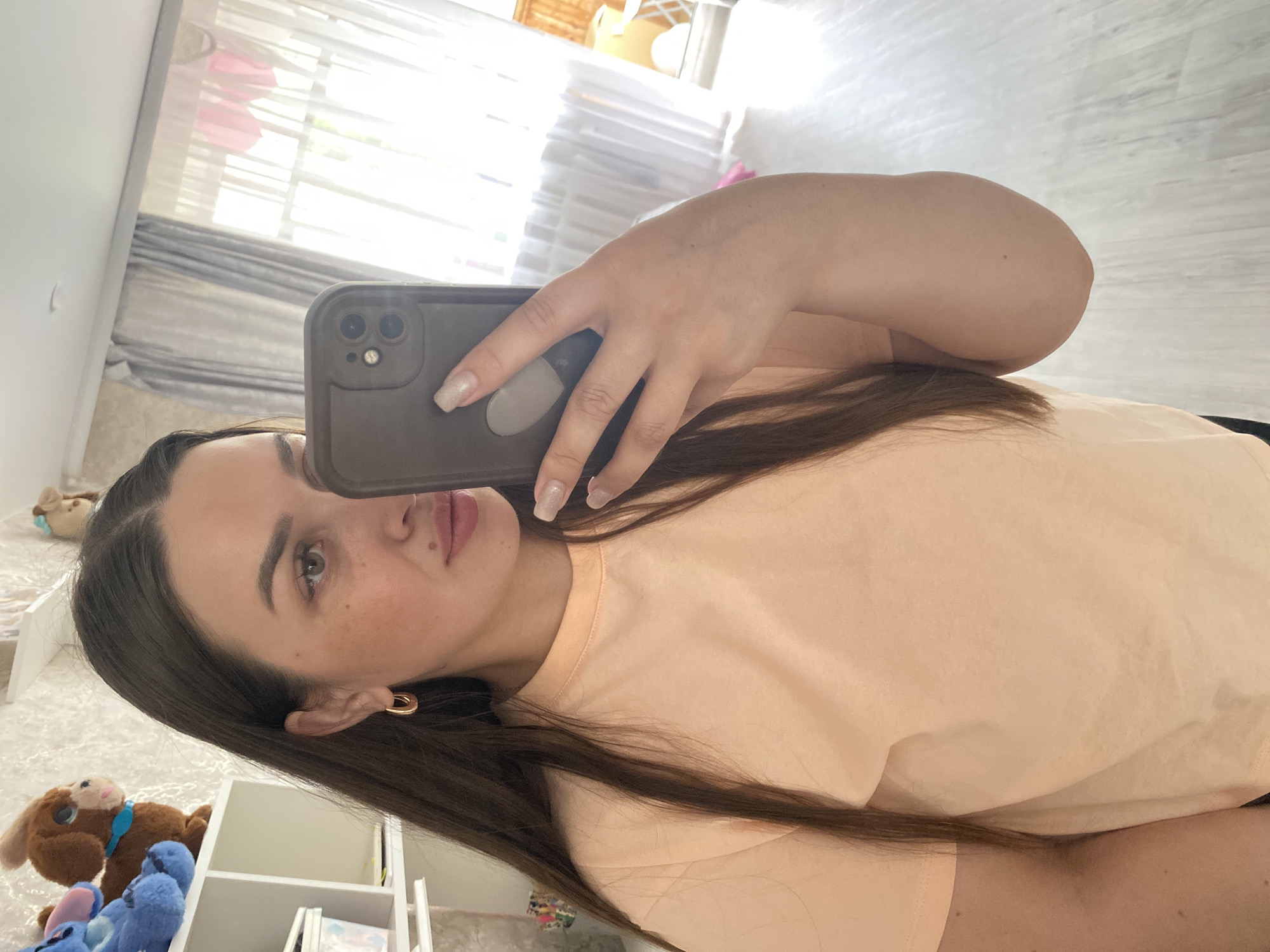 A selfie image shows a woman indoors, possibly in a bedroom. She holds a cell phone with a case that covers the camera. She is wearing an orange top and has dark hair. Behind her is a window with white curtains and a set of white shelves with various stuffed animals and books on them. The location is potentially in Chaikovsky, Russia.