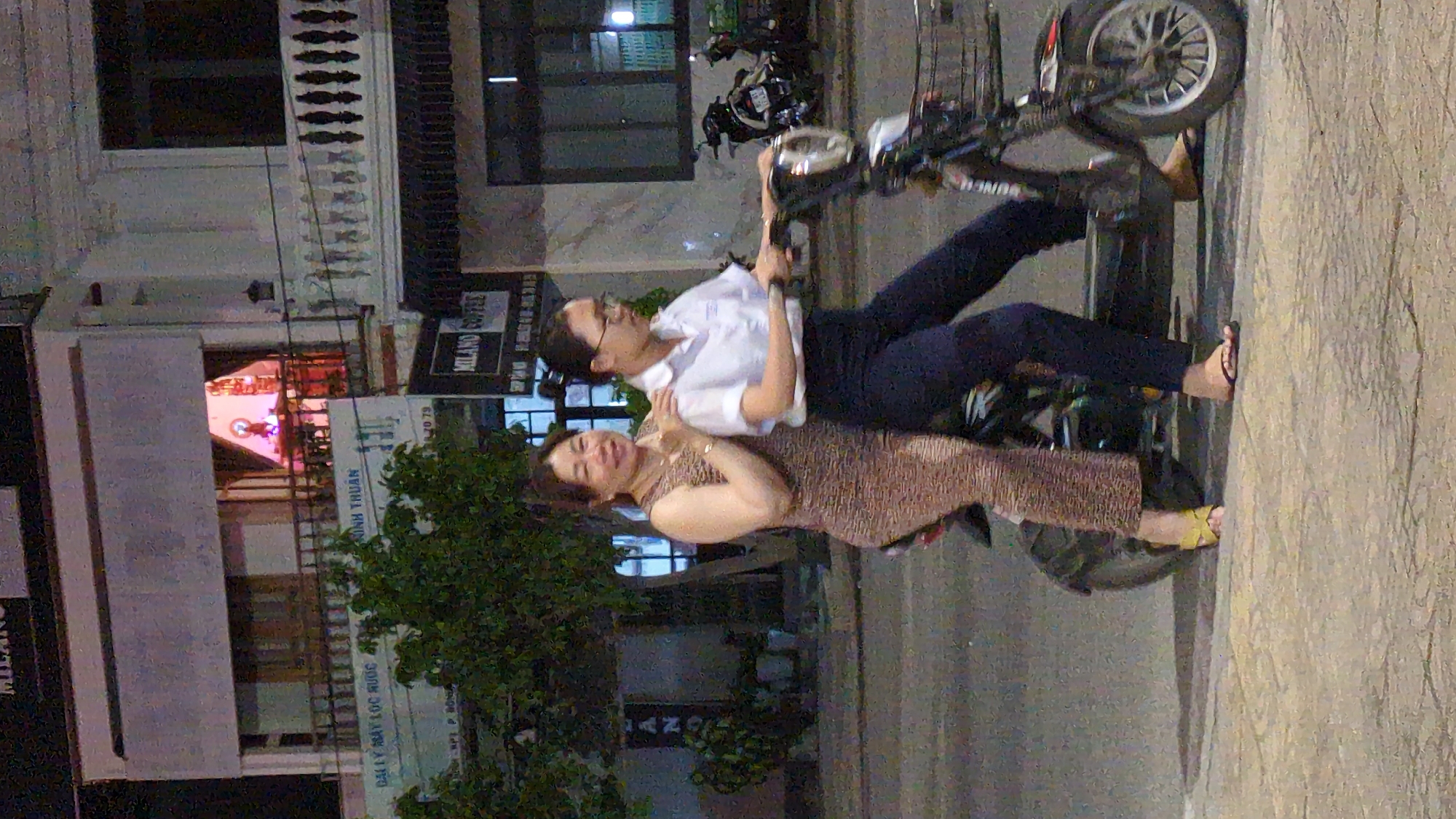 Two women are on a motorbike on a street in Phan Rang – Tháp Chàm, Vietnam. One woman is driving; she is wearing a white shirt, dark pants, and glasses. The other woman is behind her, wearing a patterned dress and holding onto the driver. The motorbike is parked on the street. Behind them is a building with a store sign on the building: "Milano Coffee." There are trees in front of the building.