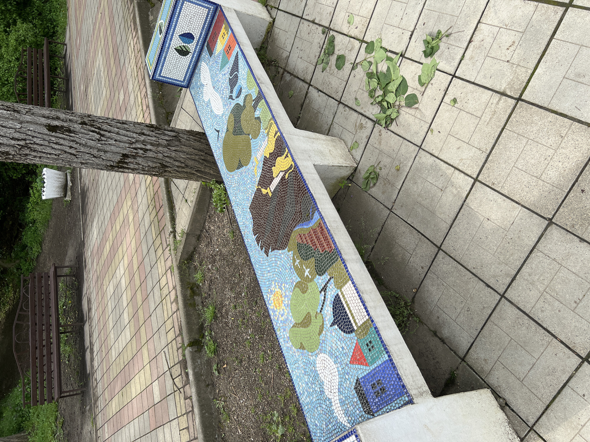 A mosaic-decorated bench stands in a park-like setting in Goryachiy Klyuch, Russia. The bench runs along a paved walkway composed of large, gray, square tiles. Green leaves are scattered on the walkway near the bench. The bench is decorated with a mosaic design in shades of blue, green, yellow, and brown. Behind the bench is a strip of bare earth, and behind that, a walkway paved with multicolored bricks. A tree trunk is positioned near one end of the bench. In the background, a dark-colored metal park bench is visible. A small, white, ceramic pot is attached to the tree trunk.