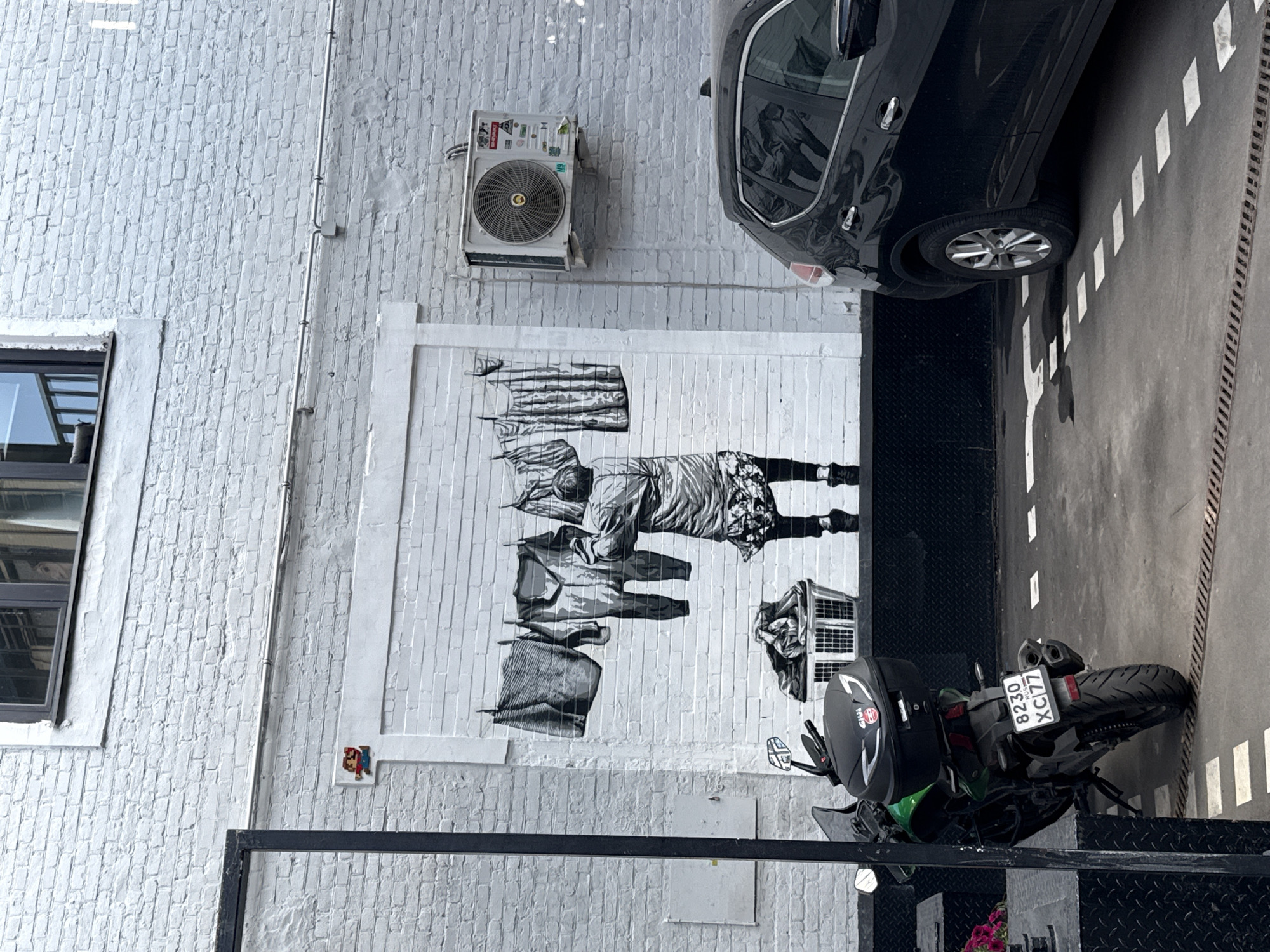 The image depicts a street art mural on a white brick wall in Moscow, Russia. The mural features a grayscale illustration of a person hanging laundry on a clothesline. Below the clothesline is a basket filled with clothes. To the left of the mural, a black motorcycle with a green body is parked. To the right of the mural, a black SUV is parked. The wall also has an air conditioning unit mounted on it, and a window above the mural. The ground is paved with asphalt and marked with white lines, suggesting it's a parking area. A small pixelated figure of Mario is visible on the wall to the left of the mural.
