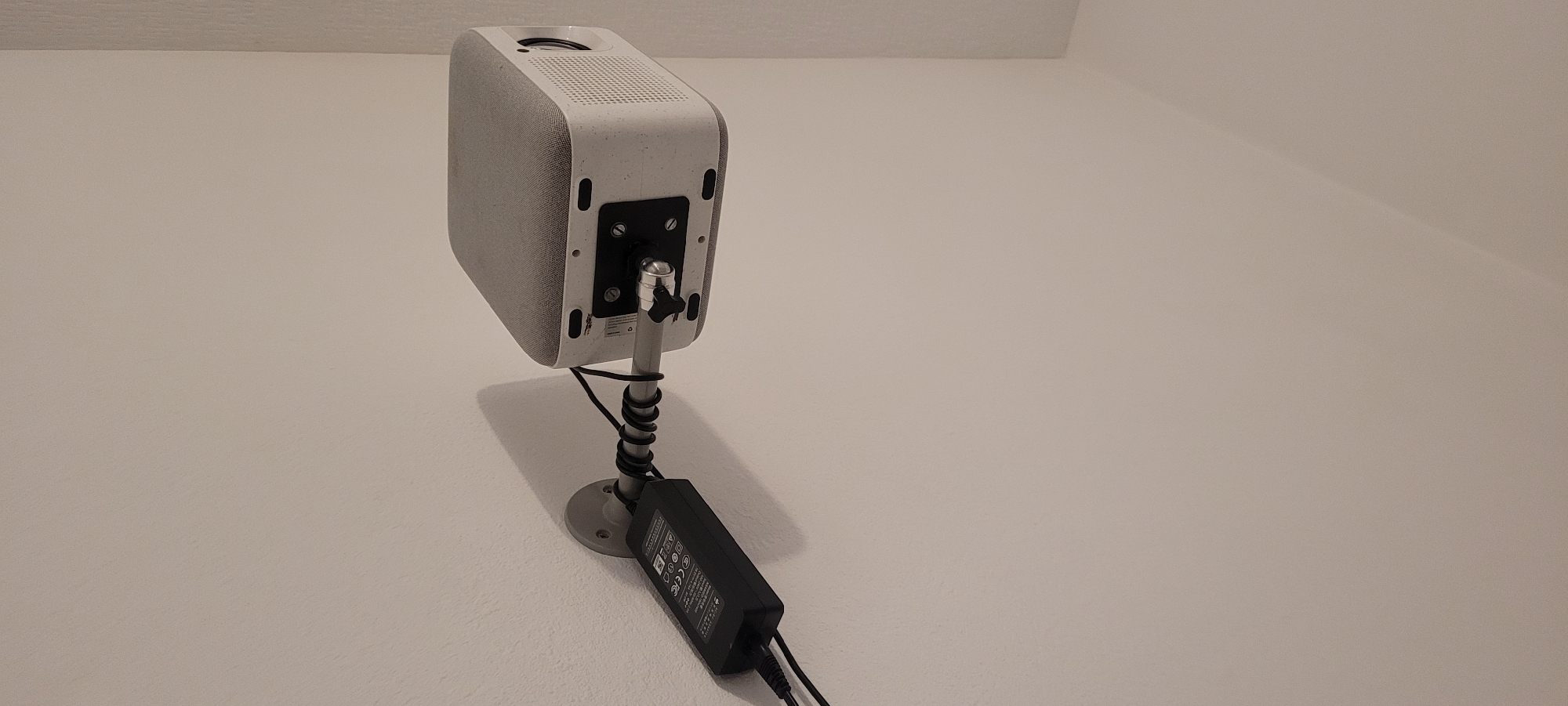 In a room located in Heusenstamm, Germany, a white and light grey rectangular projector, featuring a textile-covered top surface, is mounted horizontally on a light-colored wall. The projector displays a lens on its right side and a ventilated grille. A black rectangular mounting plate is secured to the underside of the projector with four visible screws. This plate connects to a silver-grey cylindrical arm via a pivoting joint, which is tightened by a black knurled screw. The cylindrical arm extends horizontally from a circular wall plate, which is affixed to the wall.
A black power adapter brick is positioned horizontally and secured to the cylindrical arm with multiple black spiral cable wraps. White regulatory markings, including "CE" and a crossed-out wheeled bin symbol, are visible on the adapter's top surface. Black cables emerge from the power adapter; one cable extends to the projector's power input, and another cable descends out of the frame towards the left. The wall is painted a uniform light beige or off-white color, with a subtle textured finish. A light-colored wall corner is visible on the right side of the image. No individuals are present in the scene.
