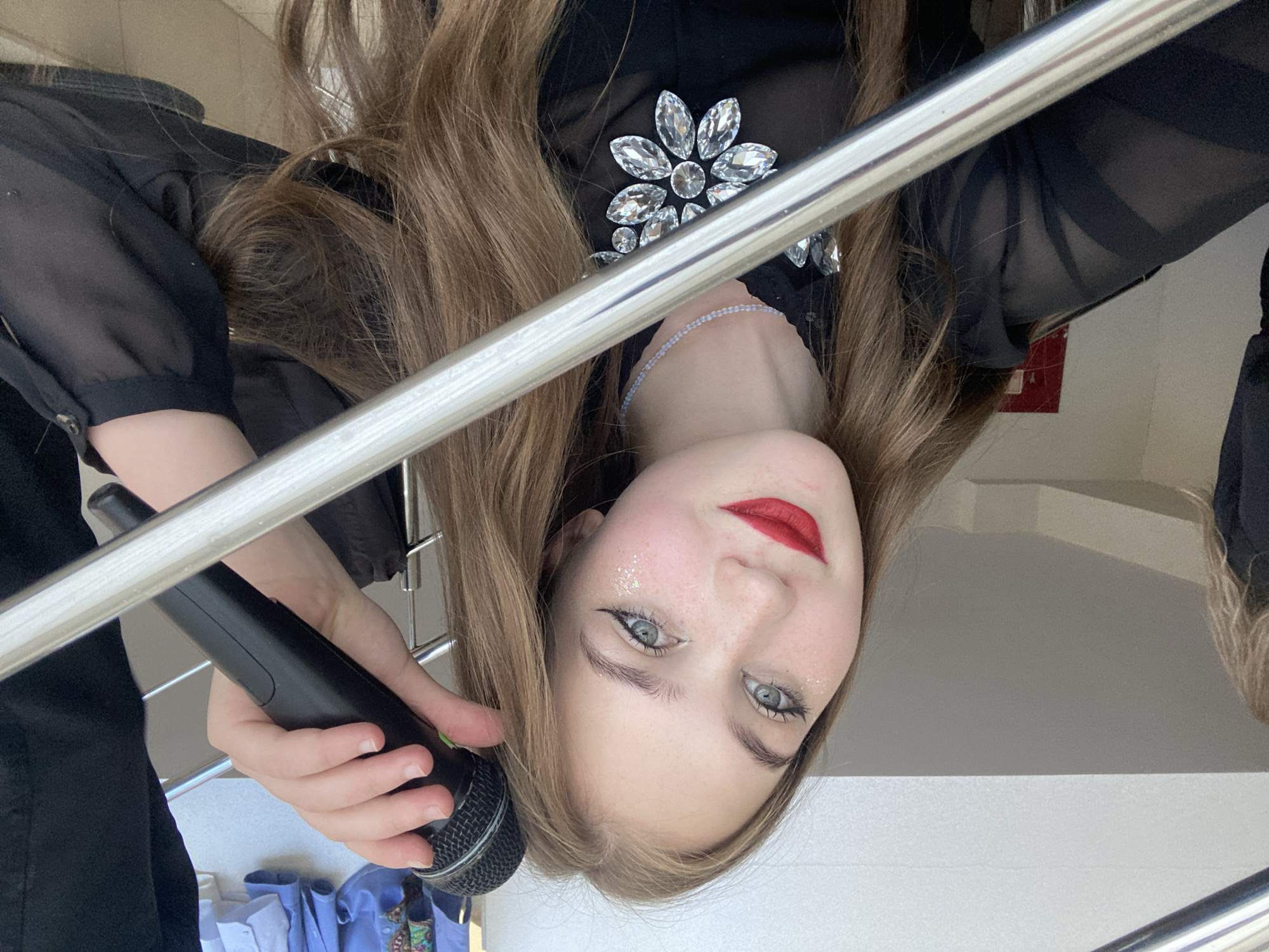 A young woman with long brown hair and light-colored eyes, wearing a black sheer top adorned with large rhinestones and a thin beaded necklace, poses for a selfie. She has bright red lipstick and glitter on her cheeks. She is holding a black microphone up to her right ear with her right hand. The background includes a metal railing, a light-colored wall, and what appears to be clothing hanging in the upper right corner. The scene is indoors, possibly in a building with stairs or a balcony due to the railing. There is no information in the image to indicate the specific location in the city of Прикамский, Russia.