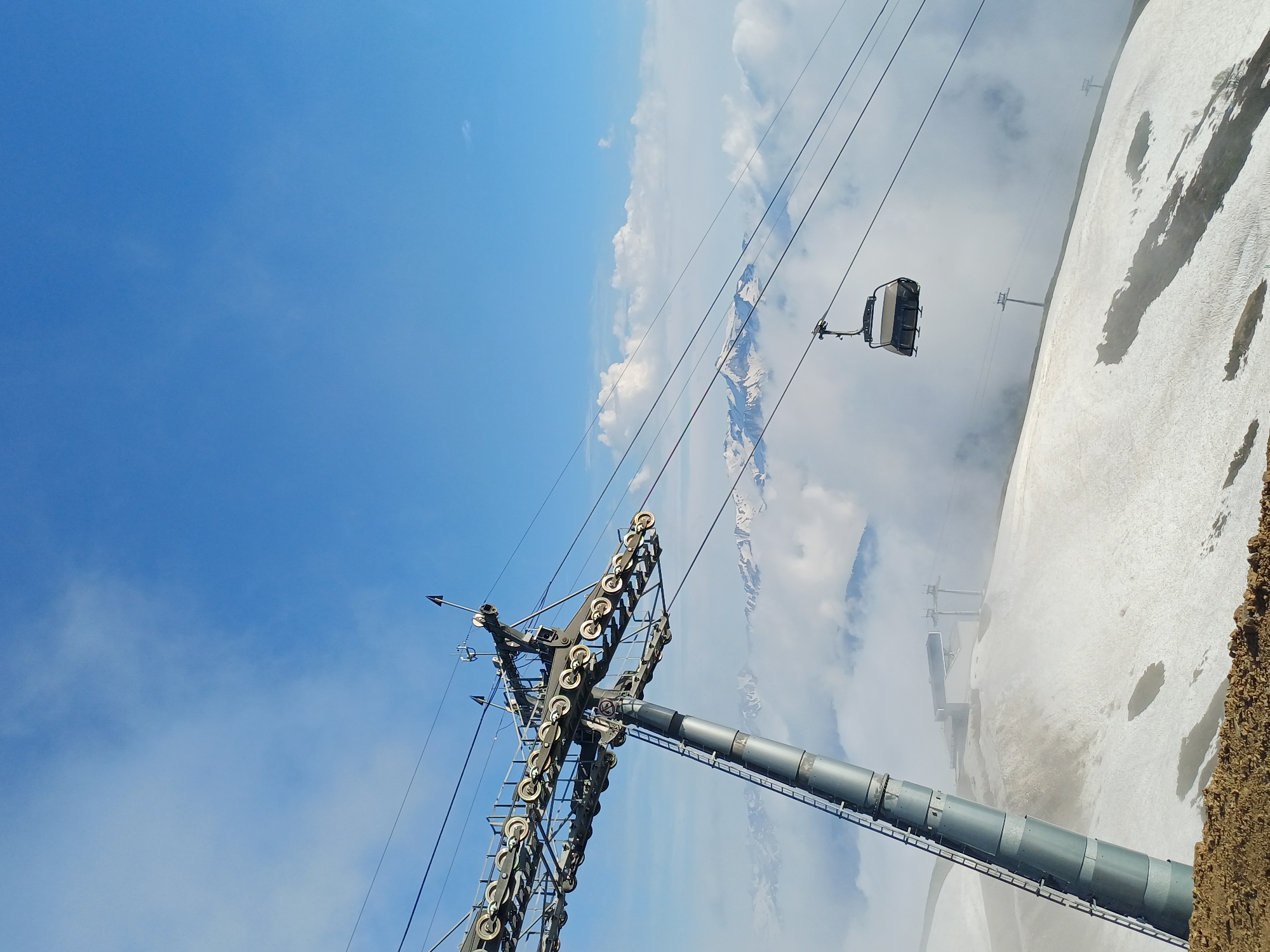 A cable car transports passengers up a snowy mountain against a bright blue sky with scattered clouds. The cable car, a modern enclosed cabin, hangs suspended from a thick cable, moving along a ski lift system. In the foreground, a large metal pylon of the ski lift system dominates the lower left portion of the frame. This pylon is a complex structure with numerous wheels and cables, indicating its function in supporting and guiding the ski lift. The mountain itself is partially covered in snow, with patches of bare earth visible, suggesting a transition between seasons or varying snow accumulation. In the distance, snow-capped mountains rise, partially obscured by clouds, adding depth to the scenic panorama. The overall impression is one of a clear, sunny day in a mountainous ski resort. The scene is likely taking place in the daytime, given the bright sunlight. The location is identified as Estosadok, Russia.