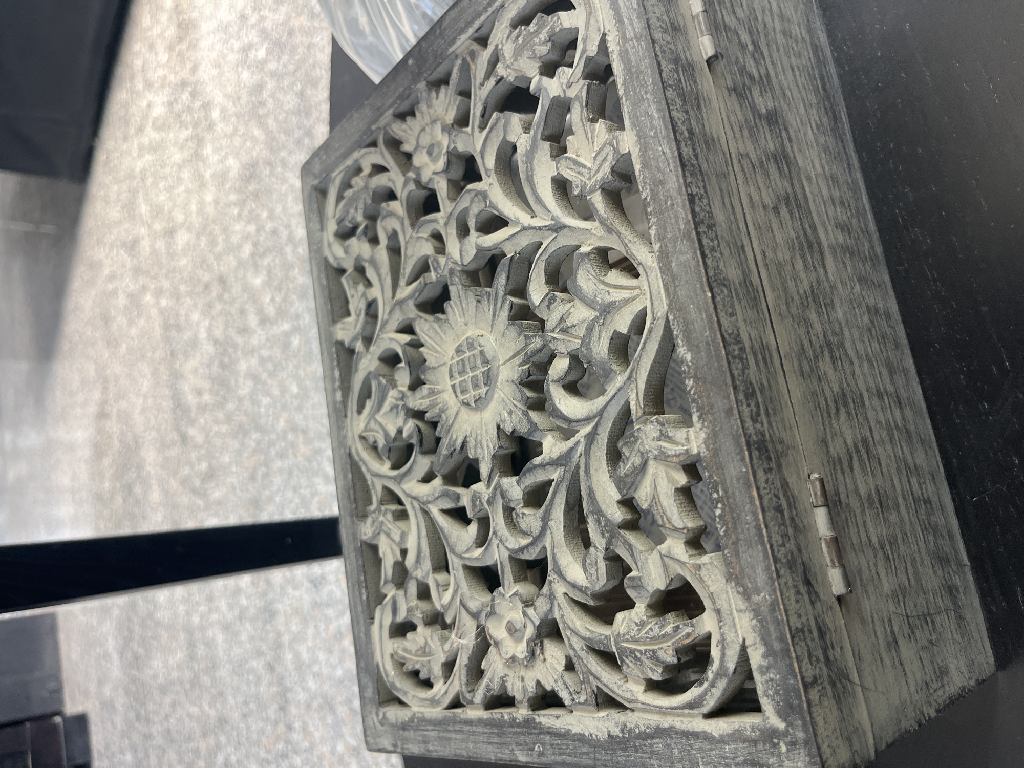 The image shows a close-up, eye-level view of a decorative wooden box with intricate carvings on its lid. The box has a distressed, weathered finish with a greyish-brown tone. The carving features floral and leafy motifs, with a prominent sunflower-like design in the center and smaller flowers and swirling vines surrounding it. The lid appears to be hinged, with visible metal hinges on the front edge of the box. The box is placed on a dark, flat surface, possibly a table or shelf. The background is blurred, suggesting a shallow depth of field, and shows a light-colored, textured surface, possibly a floor or ground, with a dark vertical object in the upper left corner. The scene is indoors, as indicated by the lighting and the indoor environment implied by the background elements. The image does not depict any people or specific actions, focusing solely on the object itself. The location of Parekklisia Community, Cyprus is not discernible from the visual information present in the image.