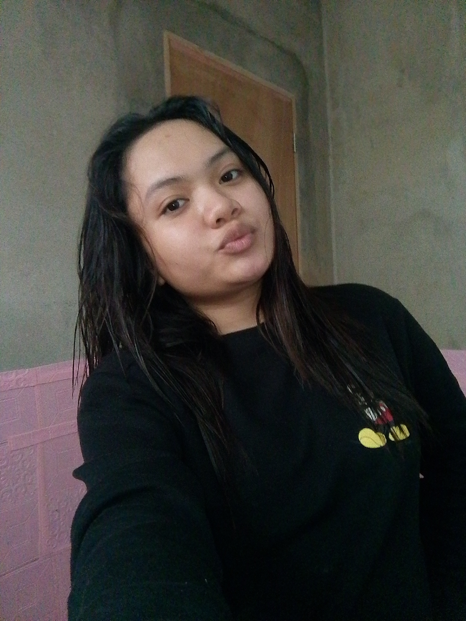 A young woman with dark hair is looking at the camera with her lips pursed. She is wearing a black shirt with a Mickey Mouse graphic on the left side of the chest. She is sitting in front of a textured gray wall and a pink couch. A wooden door is visible in the background.