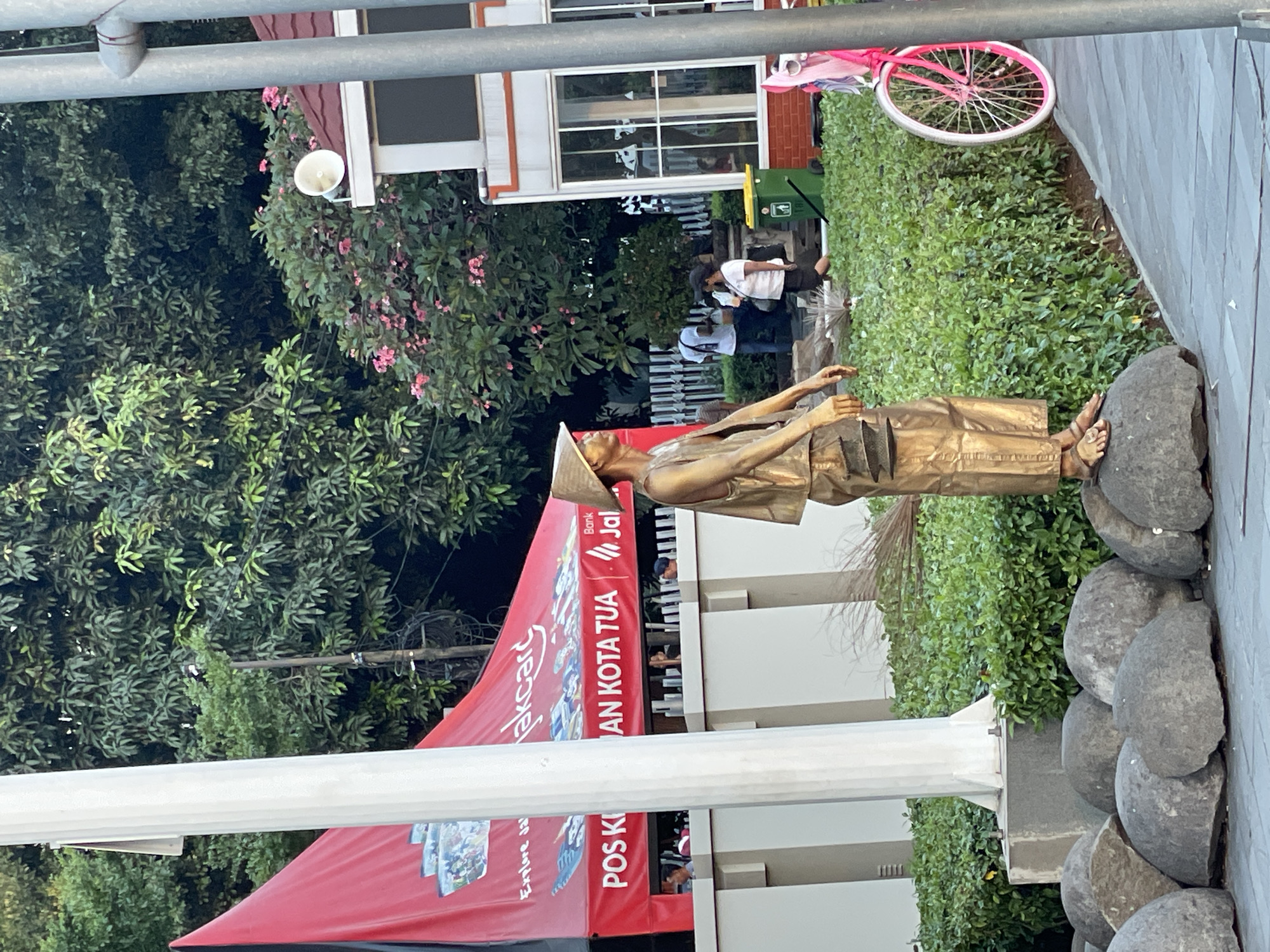 The image depicts an outdoor scene in Kota Tua, West Jakarta, Indonesia. In the foreground, a person dressed as a golden statue stands on large, rounded rocks. The statue is wearing a conical hat and traditional clothing, holding a basket-like object. A broom made of straw lies near the statue's feet. Behind the statue, a bright red tent displays advertisements for "jakcard" and "POS KOTA TUA". Several people are visible in the background, including individuals walking and standing near a building with large windows and a red roof. A pink bicycle with a white rim is parked to the right of the frame. The environment is lush with green foliage and trees, suggesting a public space or park.
