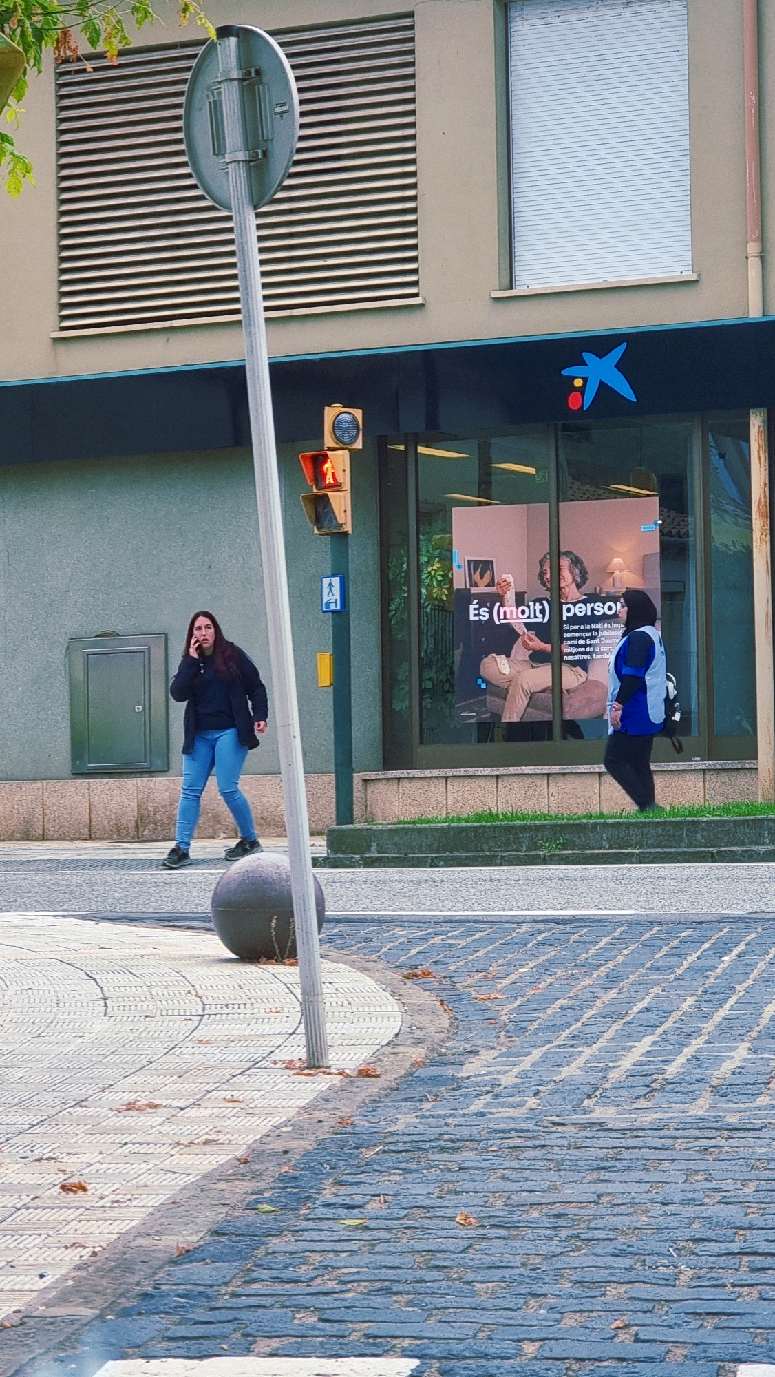 A medium shot captures a street scene in Sant Joan Les Fonts, Spain, focusing on a crosswalk and the facade of a building. In the foreground, a woman with long dark hair is talking on her cell phone, wearing a dark jacket over a navy shirt and blue jeans. She stands on a textured pedestrian walkway, a large, dark gray spherical bollard to her left. Behind her, a traffic light shows a red pedestrian signal. The building behind the woman features a large window displaying a poster with text and images of people, and a blue "la Caixa" logo is visible above the window. To the right, another person, wearing a dark uniform with a light blue vest, walks away from the camera. The street is paved with interlocking bricks in alternating shades of gray and blue, with fallen leaves scattered on the ground. The building itself has a neutral-colored facade with horizontal slatted windows and a modern entrance. A tall pole with traffic signals and signage is prominently positioned in the left half of the frame.