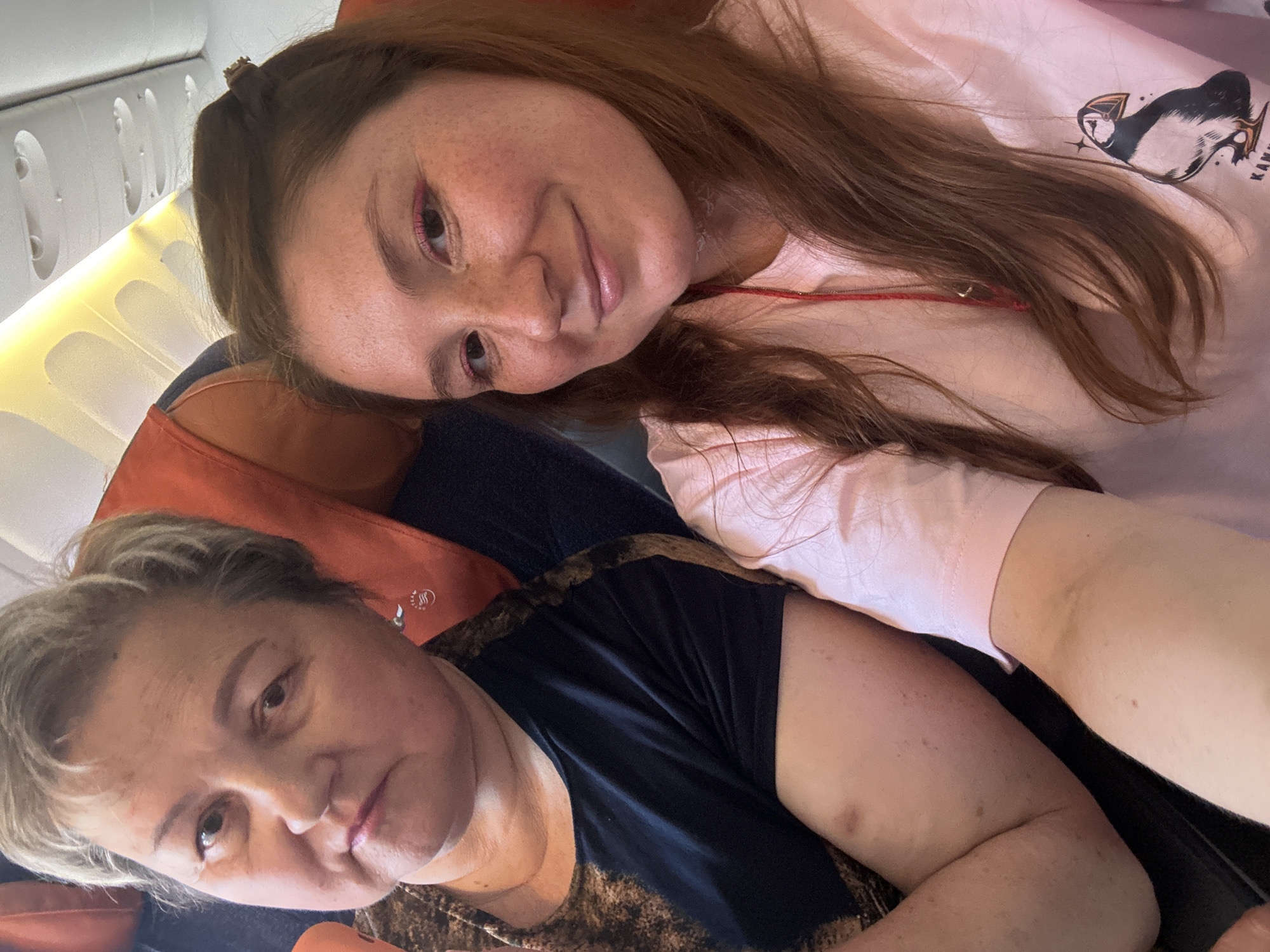 Two individuals are seated on an airplane. The person on the left is an adult female with short, graying hair and a dark, patterned top. She is looking directly at the camera with a neutral expression. The person on the right is a younger female with long, brown hair and a pink t-shirt. She is smiling and also looking at the camera. They appear to be in the same row of seats, with the airplane's interior visible in the background, including overhead compartments and lighting. The overall scene suggests travel. The location in Yelizovo, Russia is not discernible from the image.