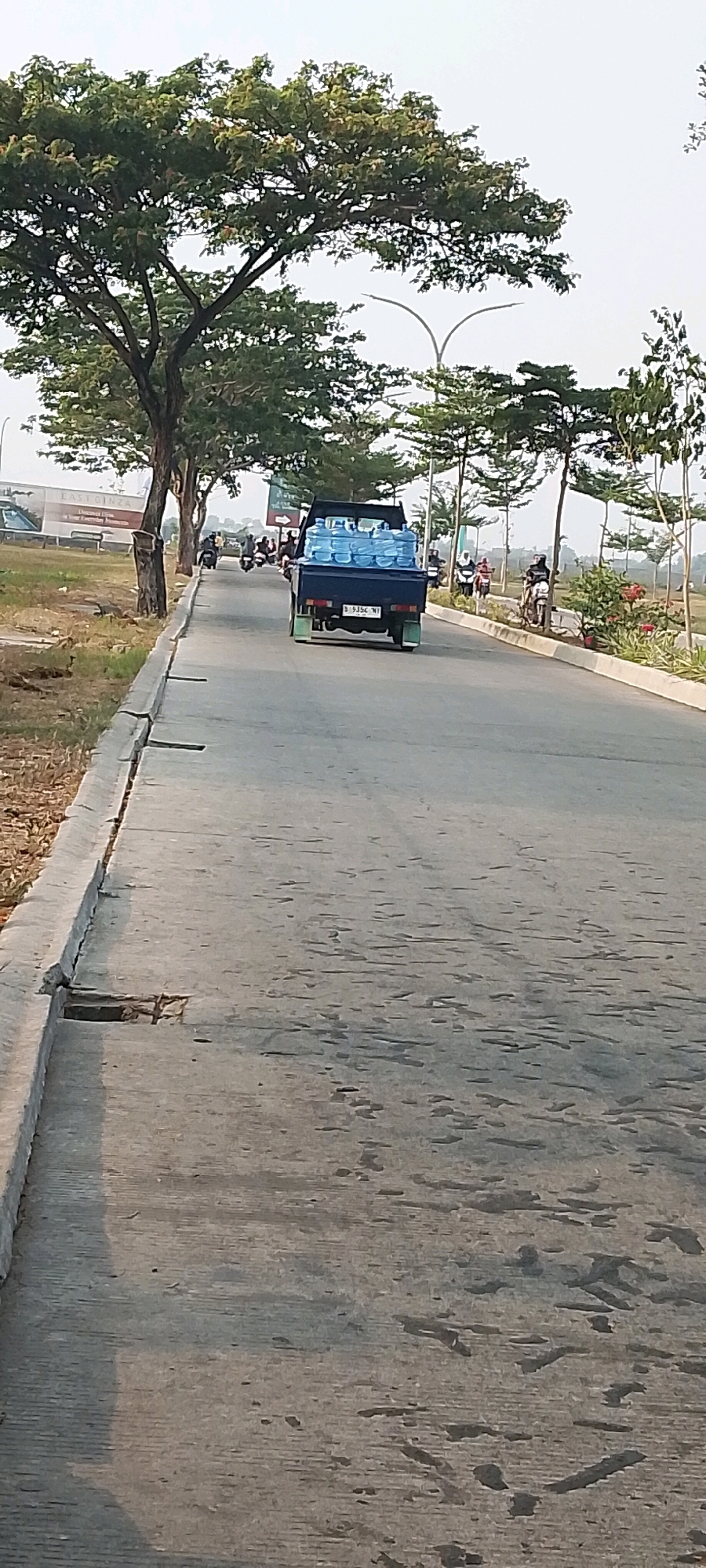 In Satriamekar, Indonesia, a dark blue pickup truck drives down a paved road, carrying several large blue water bottles in its bed. The truck has a license plate that reads "B 9354 WT". There are several motorcycles behind the truck, with riders on them. On the right side of the road, there are young trees planted along a sidewalk, and in the distance, there are more trees and buildings. The road is cracked and worn in places. The sky is bright and overcast.