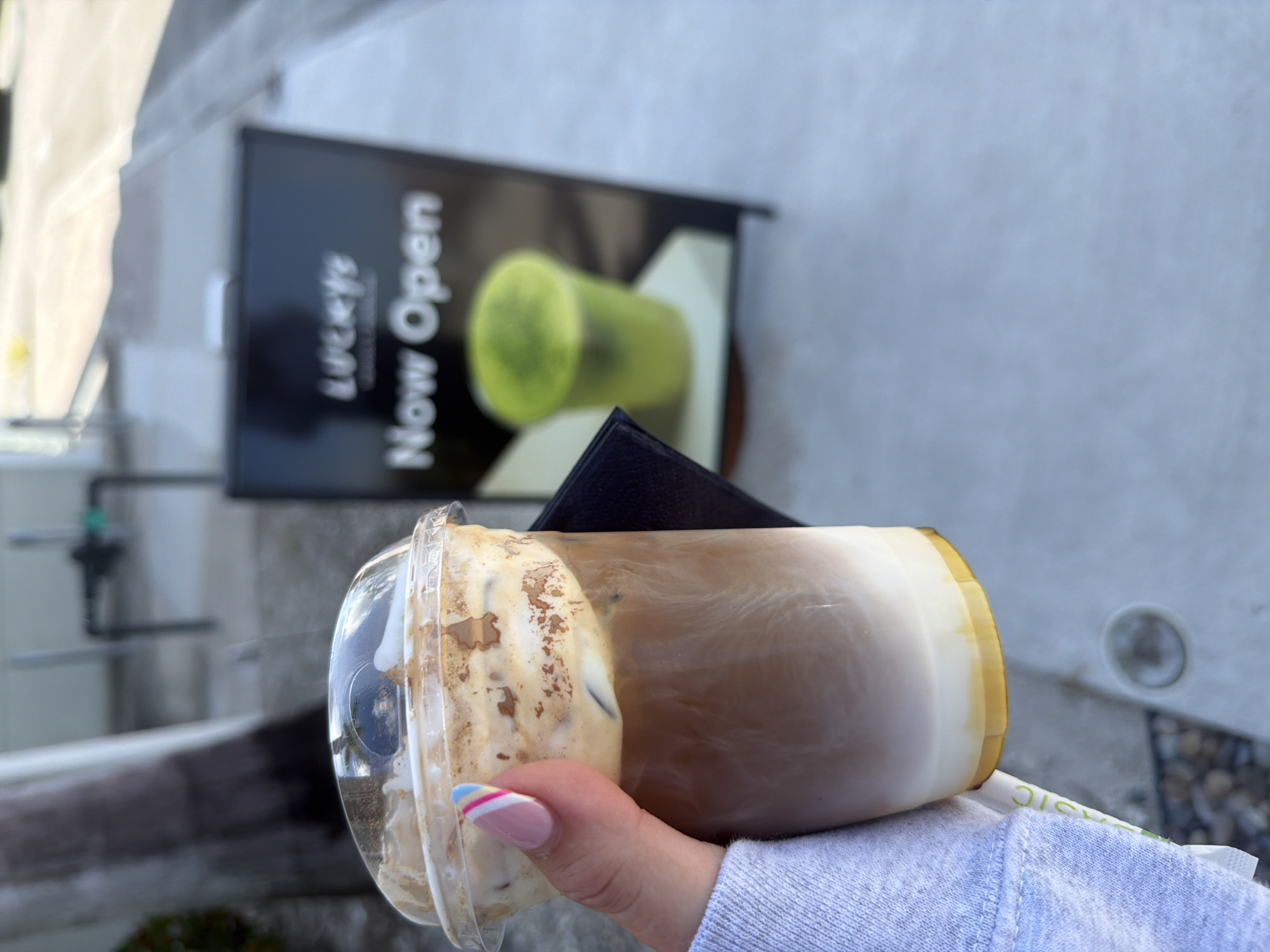 A person's hand, with manicured nails featuring pink, white, and blue stripes, holds a clear plastic cup filled with a layered iced coffee. The drink has distinct layers of white at the bottom, a caramel-colored liquid in the middle, and a foamy, creamy topping with ice cubes. The cup is partly obscured by a dark object. In the background, a black A-frame sign with "Lucky's" and "Now Open" in white lettering is visible. To the right of the sign, a blurry green beverage in a cup is also visible. The scene appears to be outdoors, on a sidewalk with a grey concrete surface, suggesting a commercial area. The lighting indicates it is daytime. This scene is likely located in North Miami Beach, United States of America, given the context of the establishment advertised on the sign.