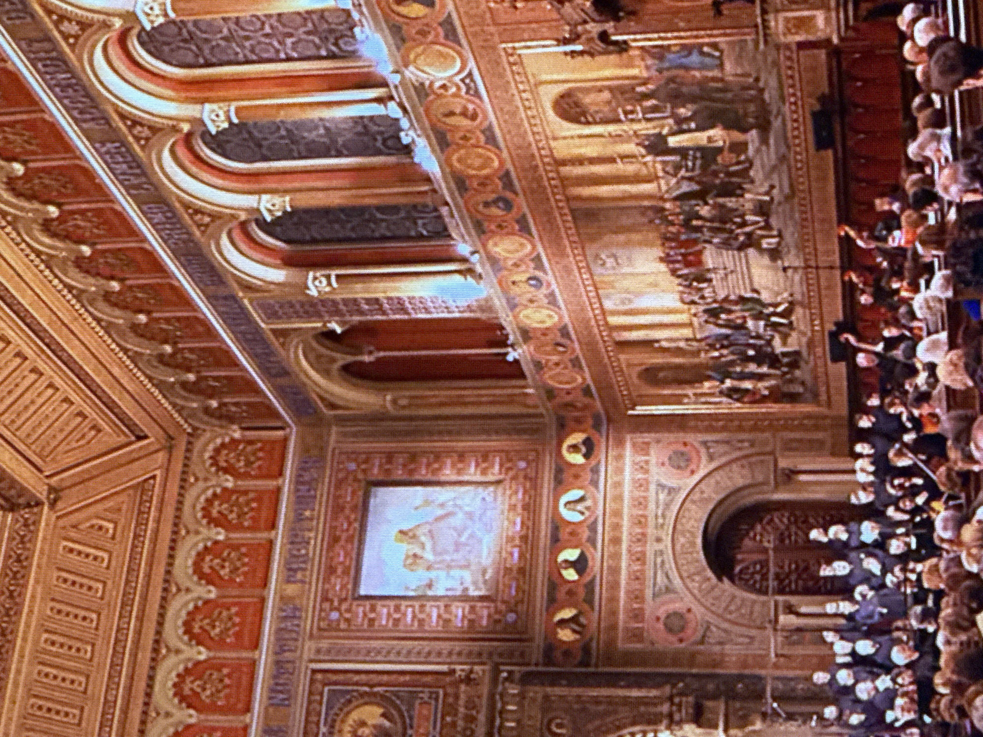 The image depicts an ornate interior, possibly a hall or theater, with elaborate architectural detailing and decorations. The walls and ceiling are adorned with rich patterns, intricate friezes, and what appear to be paintings or murals. Several arched windows or alcoves line the upper walls, some with carved details and others covered with patterned fabric or glass. Below these, a long, detailed mural spans a significant portion of the wall, depicting a scene with numerous figures in historical or ceremonial attire.
In the foreground, a large audience is seated in rows of what appear to be wooden benches or chairs. Most of the audience members are facing away from the camera, towards the mural and a stage-like area. In front of the audience, a choir or ensemble of individuals is positioned, many wearing dark robes. Some are holding sheet music, and a conductor or director appears to be present. The overall impression is of a formal gathering or performance taking place in a historically significant or artistically decorated venue. The scene is well-lit, highlighting the warm tones of the wood and decorative elements.
This image does not depict any location in Dubai, United Arab Emirates.