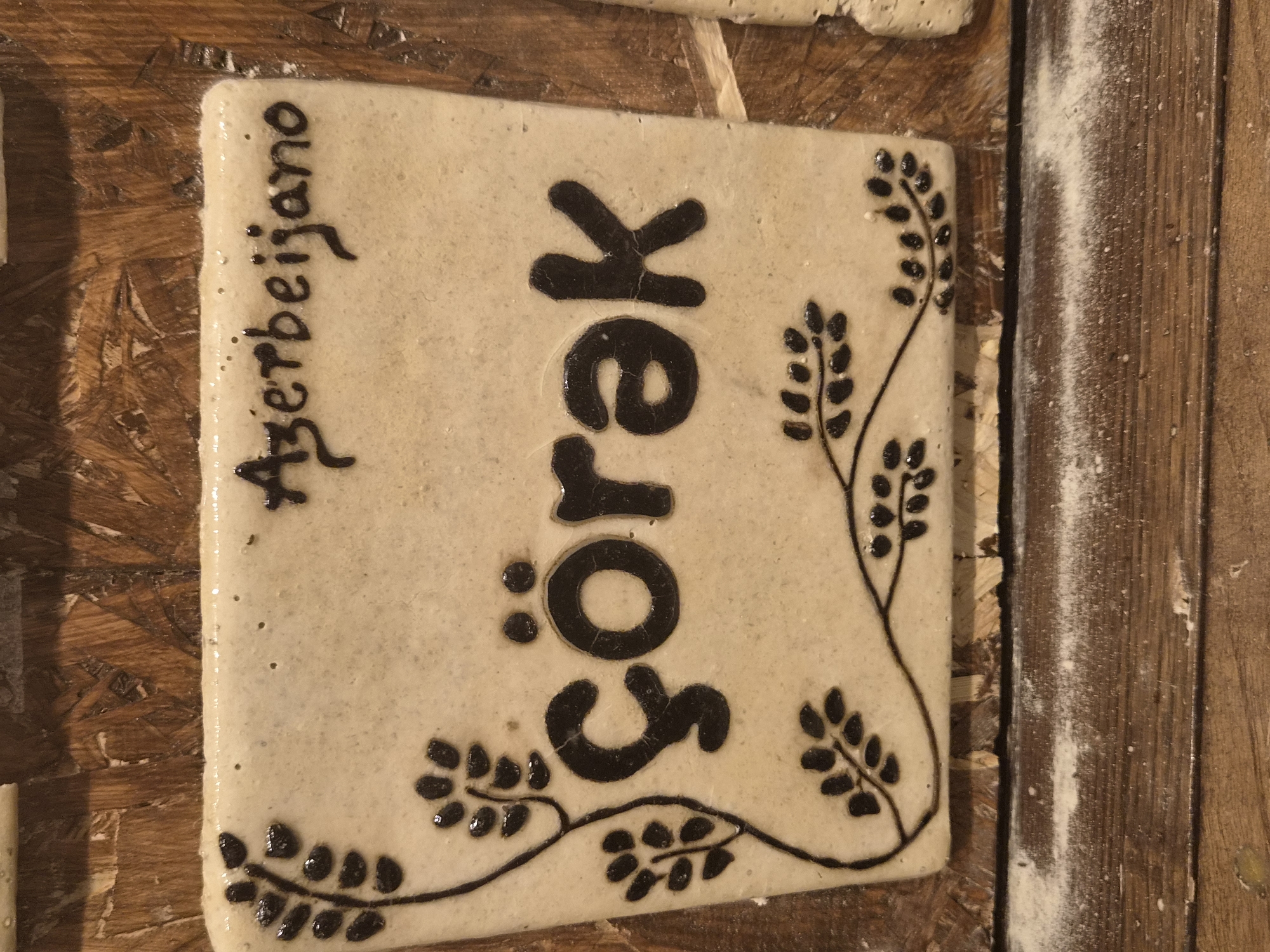 A light-colored ceramic tile with dark brown lettering and decorative motifs is placed on a wooden surface. The tile features the word "çörək" in large, stylized letters in the center, with the word "Azerbeijano" written in smaller, cursive script above it. Swirling lines with small leaf-like shapes adorn the left and bottom edges of the tile. The wooden surface appears to be a table or shelf, with visible grain and some scattered white powder, possibly flour. The overall impression is of a craft or artisanal item, possibly a decorative label or marker. This object is located in the city of Seia, Portugal.