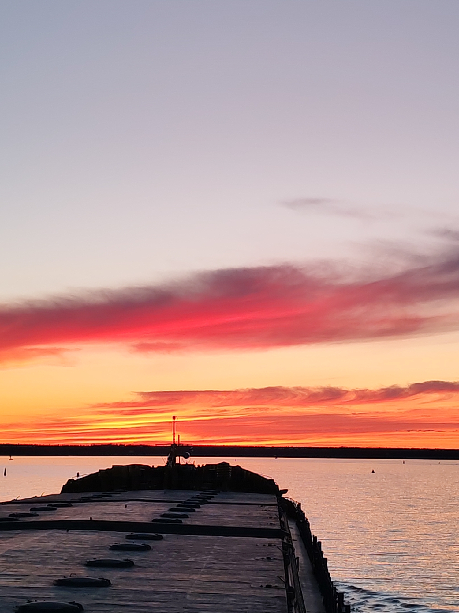 The image shows a pier extending into a body of water, likely a lake or bay, at sunset. The sky displays a gradient of colors, ranging from a light purple/gray at the top to vibrant shades of red, orange, and yellow closer to the horizon. Clouds are present in the sunset, particularly a band of bright red clouds. A dark, low-lying landmass, likely trees or shrubs, is visible on the horizon.
The pier is constructed of a dark, weathered material, possibly concrete or asphalt. It appears to have multiple access points or hatches along its surface. There is a metal pole and some structures on the end of the pier. The water surface is relatively calm, reflecting some of the colors of the sky. There are buoys or markers in the water in the distance. The location is assumed to be Topornya, Russia.