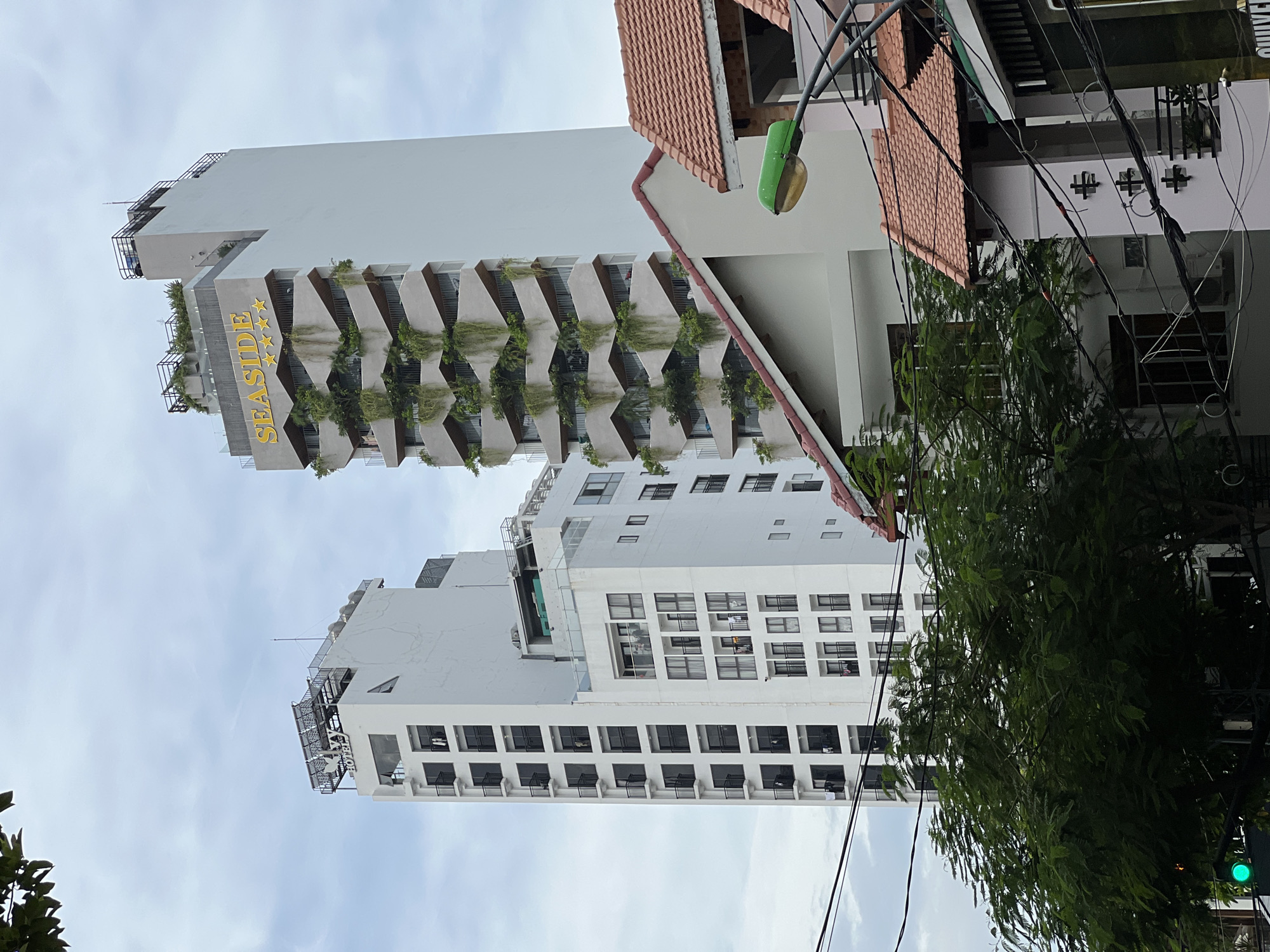 The image shows two modern high-rise buildings in Nha Trang, Vietnam, against a cloudy sky. The building on the right is taller and features a distinctive facade with angled balconies adorned with lush green plants, giving it a terraced appearance. The name "SEASIDE" is prominently displayed in yellow lettering at the top of this building, along with four yellow stars, indicating it is likely a hotel. The building on the left is also a white high-rise, with a simpler design featuring numerous balconies. Both buildings appear to be commercial or residential structures. In the foreground, there are trees and power lines, suggesting an urban environment. Partially visible in the lower right corner is a section of a building with a tiled roof and a green street lamp. The overall scene depicts a blend of contemporary architecture and the vibrant greenery characteristic of tropical cities like Nha Trang.