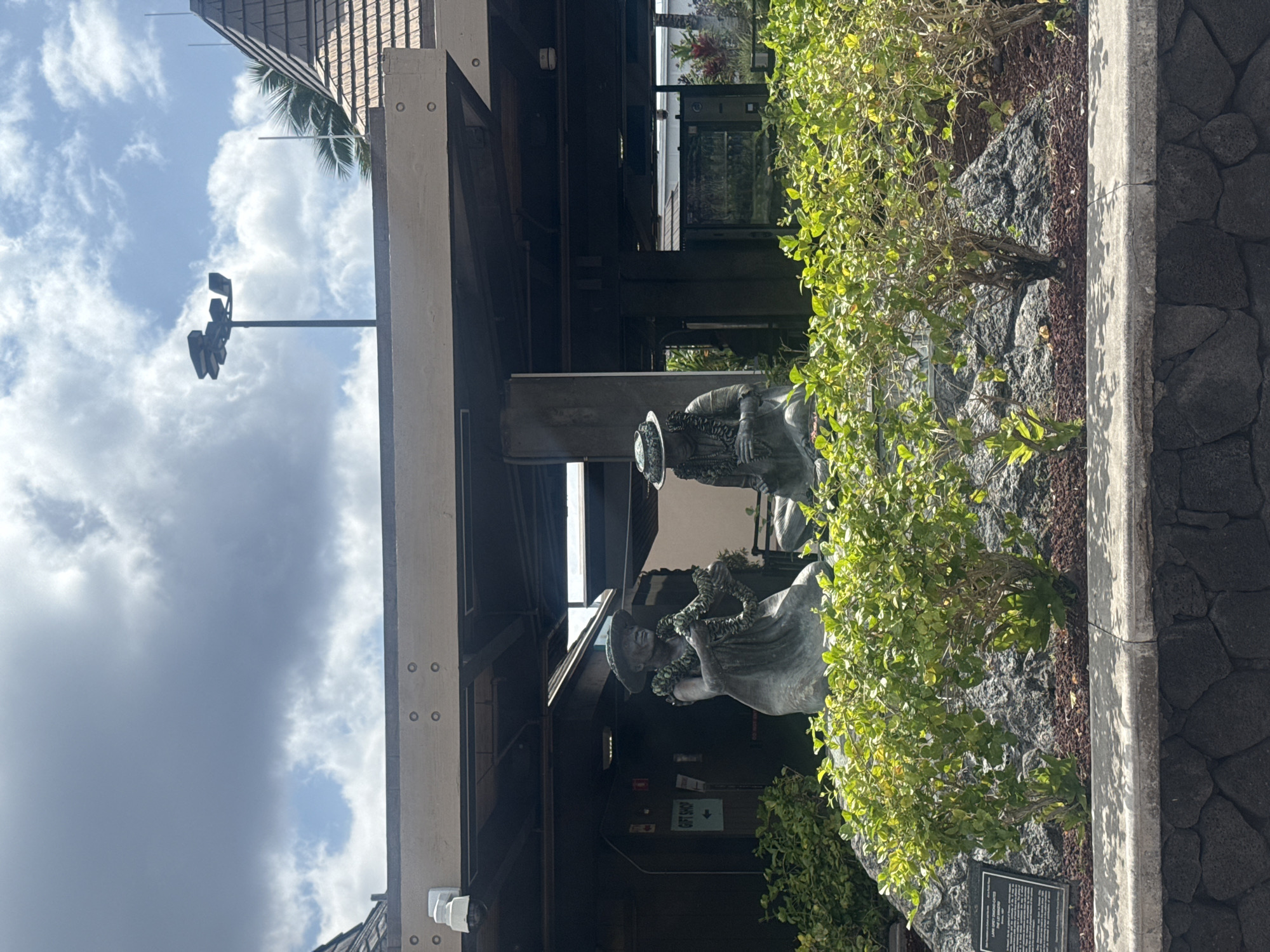 The image depicts an outdoor scene in Kailua, United States of America, under a partially cloudy sky. Two bronze statues of elderly Hawaiian women, identified as "The Lei Makers," are positioned prominently in the mid-ground. The statue on the left is seated, wearing a wide-brimmed hat and a lei around her neck, actively depicted in the process of making a lei. The statue on the right is also seated, wearing a similar hat and lei, holding materials consistent with lei making. Both sculptures rest on a base composed of dark, irregular rock formations, surrounded by vibrant green shrubs and dark brown mulch. In the immediate foreground, a retaining wall constructed from dark, irregularly shaped stones is topped with a light-colored concrete ledge. Behind the statues, a dark-colored building structure features a broad wooden overhang supported by dark pillars. A white security camera is affixed to the underside of this overhang, positioned above and to the left of the left statue. A "GIFT SHOP" sign with a directional arrow is visible on the building wall behind the left statue. The building's roof, partially visible on the right, exhibits a multi-tiered shingled design. A tall black pole equipped with multiple floodlights extends into the sky on the upper right side of the frame, and a palm tree frond is partially visible near the right edge of the roofline. Reflections on glass surfaces are visible within the building structure behind the right statue, with a faint, indistinct figure possibly visible through the glass in the background.