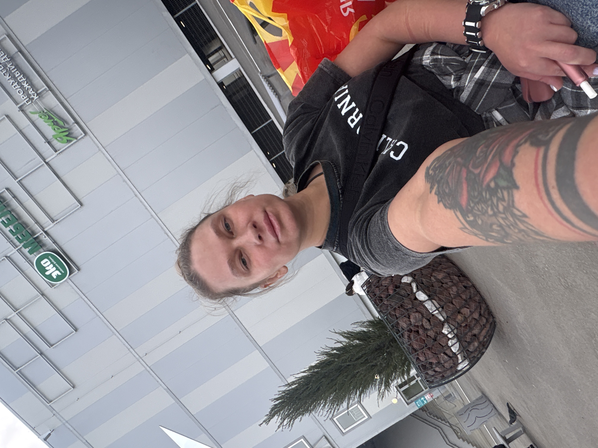 A medium, eye-level, indoor shot depicts a woman angled to the right holding what appears to be an electronic cigarette. She wears a black t-shirt, a watch, and a flannel-like top and has tattoos on her arm. In the background there is a building labeled "Semya" written in Russian. Below it it says "Eco" in English and Russian and "Products for everyone". In the lower right corner, there is a plant growing in a cage filled with rocks. From the location, the photo was likely taken in Perm, Russia.