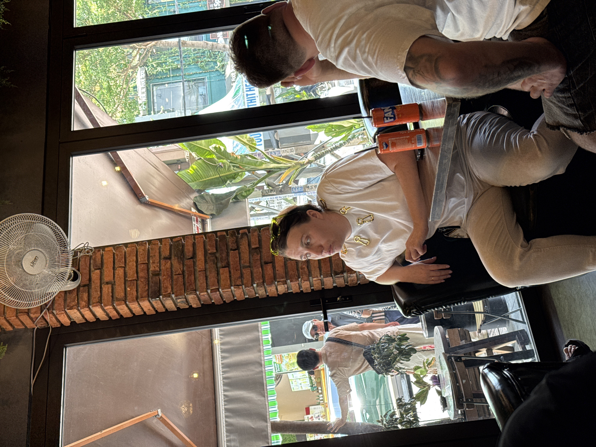 The image depicts the interior of a cafe or restaurant in Nha Trang, Vietnam, with a focus on patrons seated at a table. A woman with blonde-tinted sunglasses perched on her head and a gold chain necklace sits in the foreground, looking to her left. She wears a white t-shirt and light-colored pants. On the table in front of her are two orange cans of Fanta, one with a straw. To her right, a person with short hair and a tattoo on their arm is visible, facing away from the camera, wearing a white t-shirt and dark shorts.
Through the large glass windows behind the woman, the street scene of Nha Trang is visible. A man in a light-colored shirt with a bag slung across his shoulder stands outside, facing the window and gesturing towards it. Another man in a white hat and sunglasses stands next to him. Greenery, including large leaves and trees, is abundant outside, suggesting a lush environment. A brick pillar with a ceiling fan mounted on it is also part of the interior decor. The cafe's interior appears modern with dark seating and metal-accented tables.