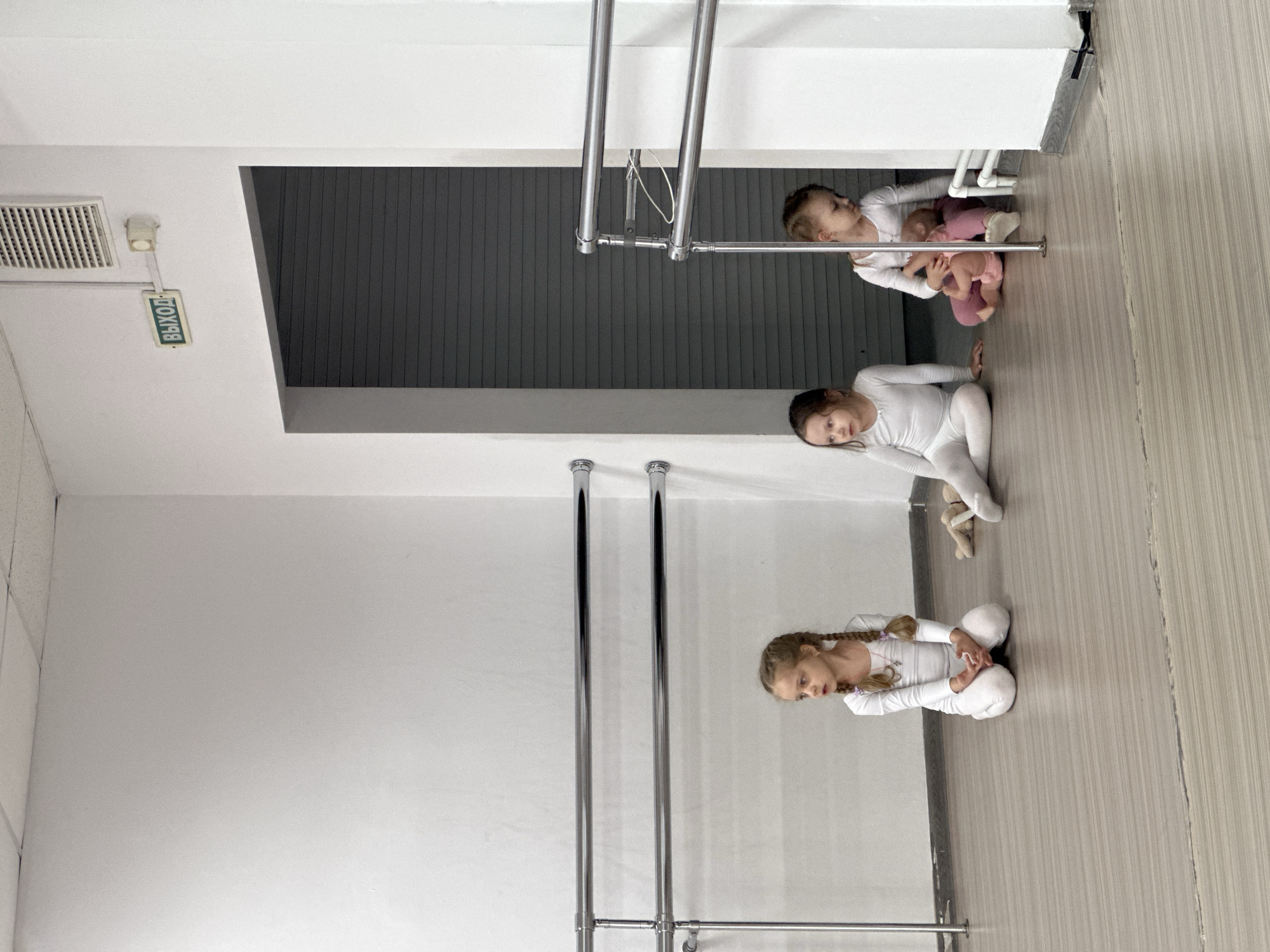 The image captures three young girls inside what appears to be a dance or ballet studio located in Votkinsk, Russia. The room has plain white walls and a light brown wood-grain laminate floor. Two horizontal chrome ballet barres are affixed to the left wall. A doorway without a door is visible on the right, leading into a darker area with horizontally grooved, possibly corrugated, walls. Above this doorway, a green illuminated sign displaying "ВЫХОД" (EXIT) in Cyrillic is mounted, accompanied by a white rectangular ventilation grate and a small white sensor unit. A white baseboard runs along the bottom of the walls, with some white pipes and a small radiator unit visible in the lower right corner.
From left to right:
1. A young girl is kneeling on the floor, dressed in a white long-sleeved leotard and white tights. Her light brown hair is styled in two braids with purple ties, and she wears a small pink necklace. She faces left with a slightly solemn expression.
2. A second young girl is seated on the floor with her legs extended, also in a white long-sleeved leotard and white tights. She faces forward. A pair of light brown ballet slippers are on the floor near her left foot.
3. A third young girl is seated on the floor near the doorway, holding a pink baby doll in her lap. She wears a white long-sleeved top and pink pants. Her gaze is directed towards the left. A vertical chrome pole is visible in front of her.
The girls are not actively interacting with each other but are seated or kneeling within the practice space, appearing to be at rest or awaiting instruction.