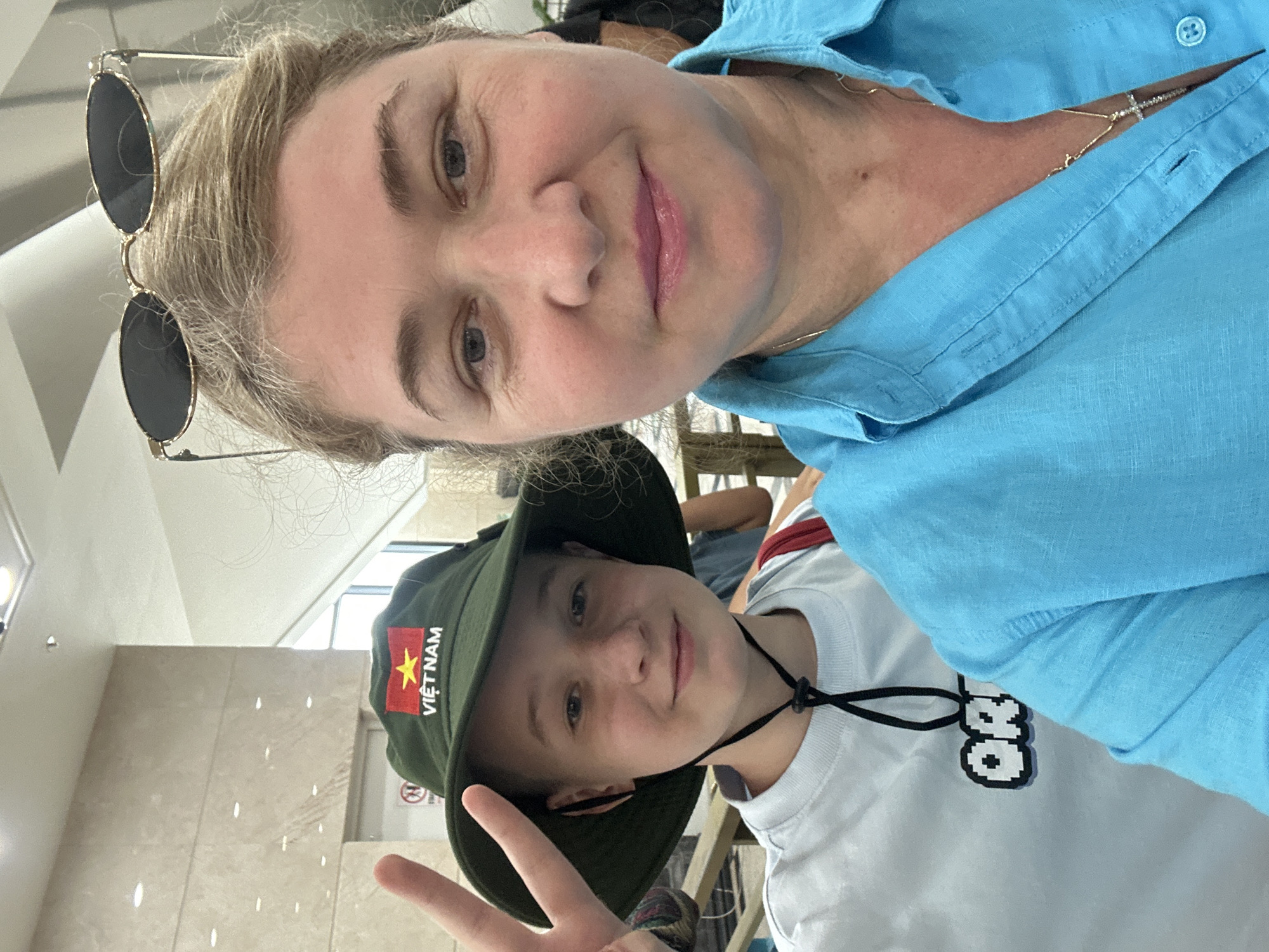 A selfie shows a woman and a child wearing a hat with a Vietnam flag patch. The woman is in the upper portion of the frame, wearing a blue button-down shirt and sunglasses perched atop her head. She is looking at the camera and smiling. Below her, the child is also smiling and making a peace sign with their hand. They are wearing a light-colored T-shirt with a pixelated design. The location appears to be indoors, with a tiled wall visible in the background. Based on the flag patch on the hat, the photo was taken in Vietnam. Based on the request, the photo was likely taken in the city of Nha Trang Ward, Vietnam.