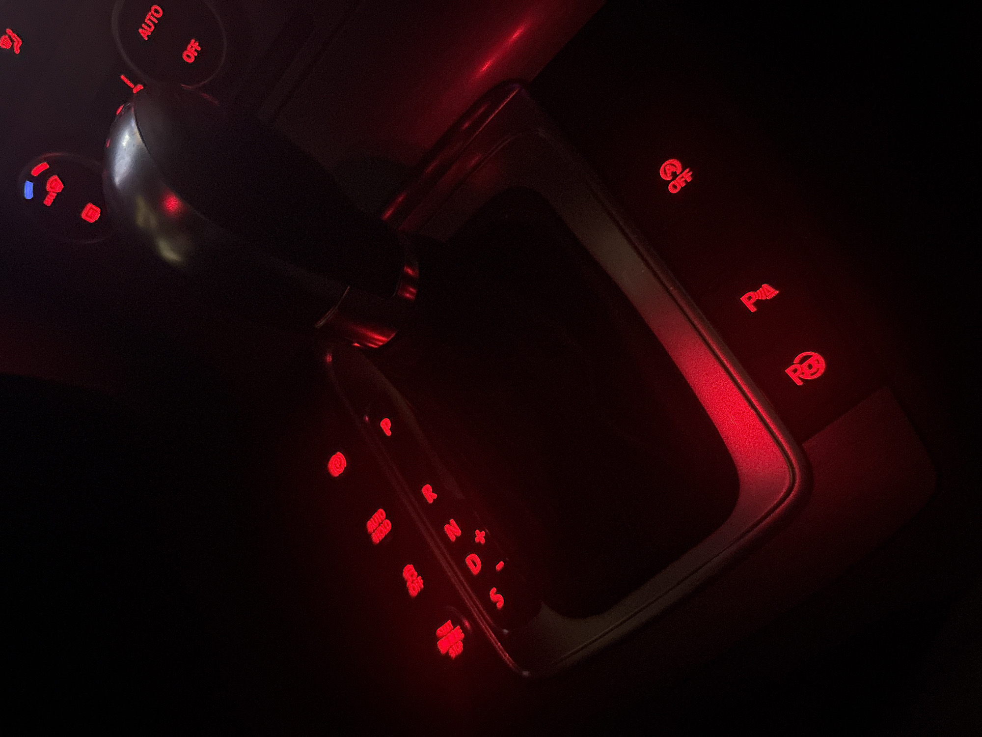 This is a close-up, low-light photograph of a car's automatic gear shift and surrounding control buttons, illuminated by red backlighting. The dominant object is the gear shift lever, a metallic and black plastic component, positioned in the center of the frame. To the left of the gear shift, several buttons are visible, some with red illumination and others with blue. These include buttons labeled "AUTO," "OFF," and what appears to be a traction control button.
To the right of the gear shift, a metallic trim piece outlines the gear selector area. Further to the right, more illuminated red buttons are visible, including one with the symbol "P" for Park, and another with a parking sensor symbol. Along the gear shift itself, the typical selection indicators for an automatic transmission are visible and illuminated red: "P," "R" (Reverse), "N" (Neutral), "D" (Drive), and "S" (Sport). There are also plus and minus symbols, indicating manual gear selection.
The overall scene is dark, suggesting it is nighttime or the interior lights of the car are predominantly on. The red lighting creates a mood that is both functional and perhaps a bit dramatic. There are no people visible in the image. The context of Burgas, Bulgaria, does not provide specific visual cues within the image itself, but situates the photograph within a particular geographical location. The image focuses solely on the interior controls of a vehicle.