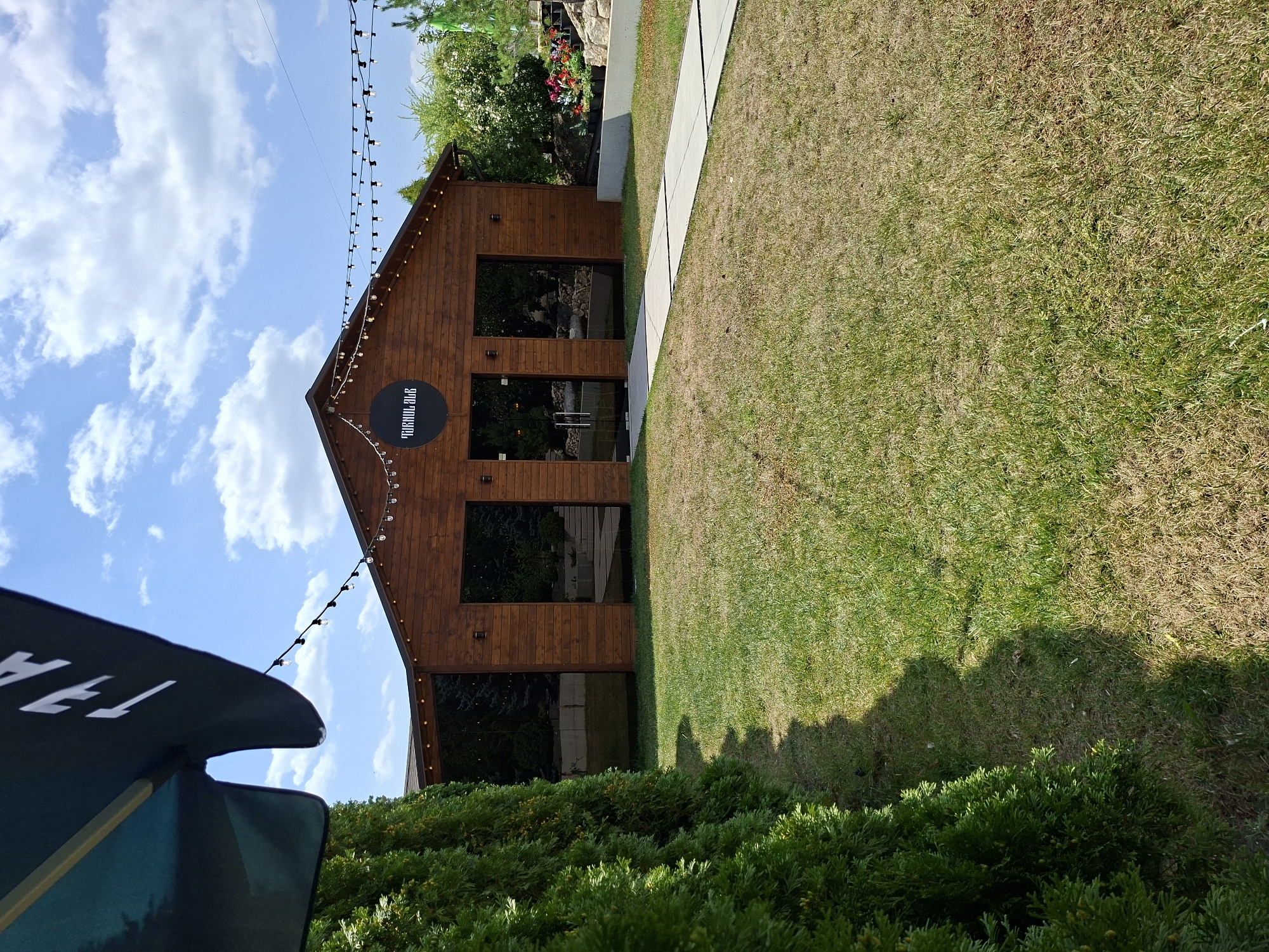A modern wooden building with a dark circular sign bearing white Cyrillic text is situated in a grassy outdoor area. The building has large glass doors and windows, and is adorned with string lights along its eaves. A paved walkway leads to the entrance, and a dense green hedge runs along the left side of the image. The scene appears to be a commercial establishment, possibly a cafe or restaurant, located in Costești, Moldova, under a bright, cloudy sky.