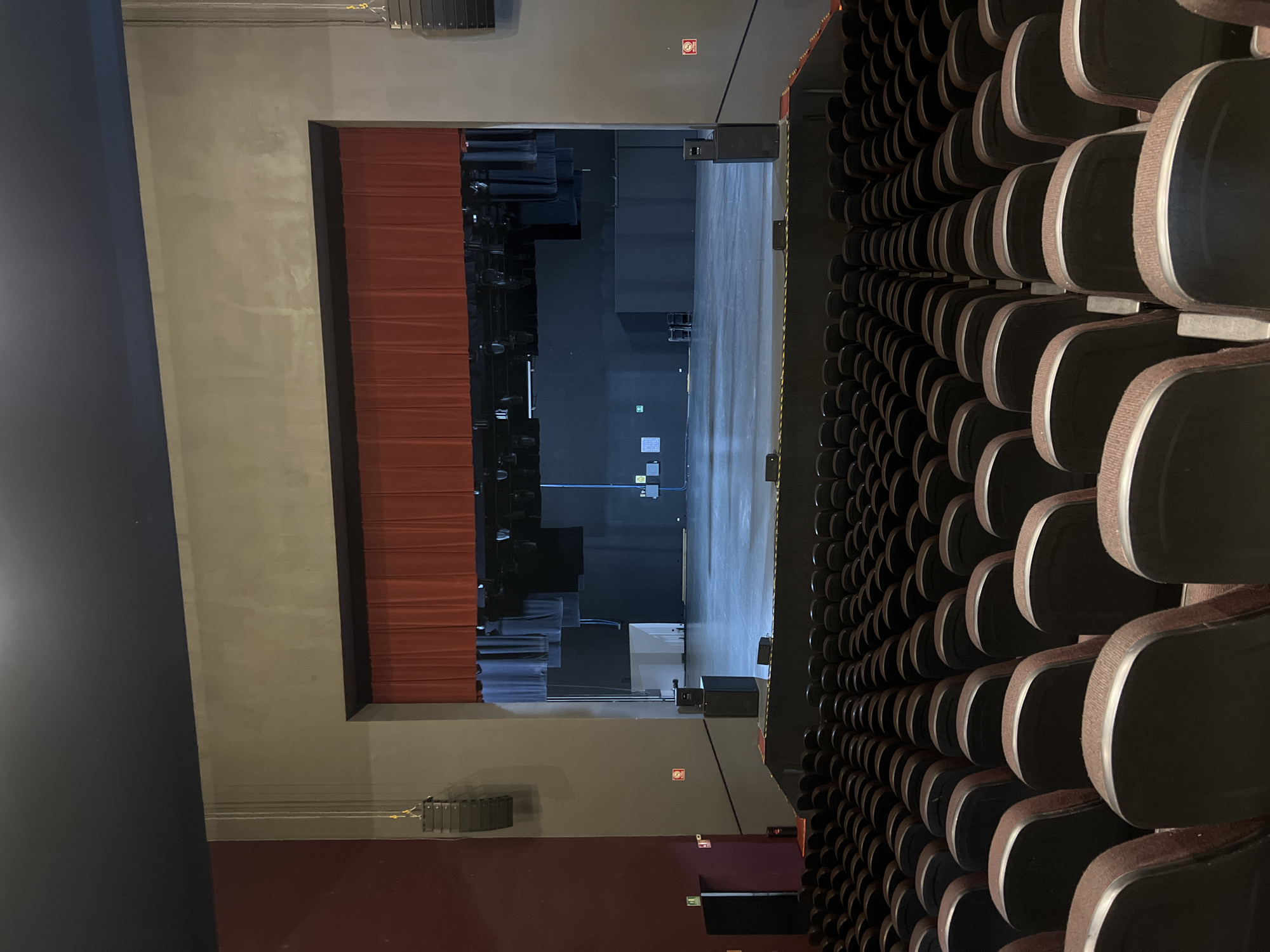 The image depicts an empty auditorium or theater, viewed from an elevated position looking towards the stage. Numerous rows of unoccupied, dark-colored upholstered seats with light brown or beige trim line the right side of the frame, receding into the background.
To the left, a wide stage is partially obscured by a deep red, pleated curtain on the left side of the stage opening. The stage floor is a light, reflective surface, while the back and sides of the stage area are dark, suggesting black backdrops or walls. Various pieces of stage equipment, including what appear to be speaker arrays and lighting rigs, are visible in the dimly lit stage area.
The overall lighting is subdued, suggesting the venue is not currently in use or being prepared for an event. There are no people visible, indicating no activity is taking place. Notable details include multiple red square signs with white emergency symbols (likely fire safety indicators) affixed to the walls, and a green square sign with a white arrow indicating an exit on the stage's rear wall. The setting is an indoor venue in Puebla, Mexico.