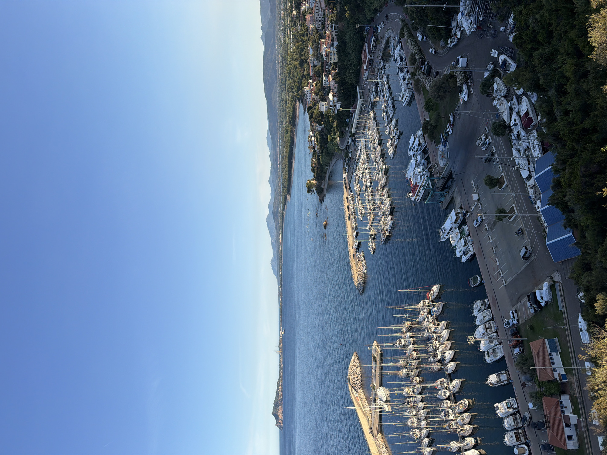 The image shows an aerial view of a harbor in Santa Maria Navarrese, Italy, filled with numerous sailboats and motorboats docked in the water. The harbor is protected by two stone breakwaters. Beyond the harbor, a coastal town with white and terracotta-roofed buildings is visible, nestled amongst lush green trees. In the background, a range of mountains can be seen under a clear blue sky. A road winds through the town, with several cars parked along it and in parking lots. A few buildings, possibly boat maintenance facilities, are situated near the shore. The overall scene depicts a serene coastal landscape with a thriving maritime activity.
