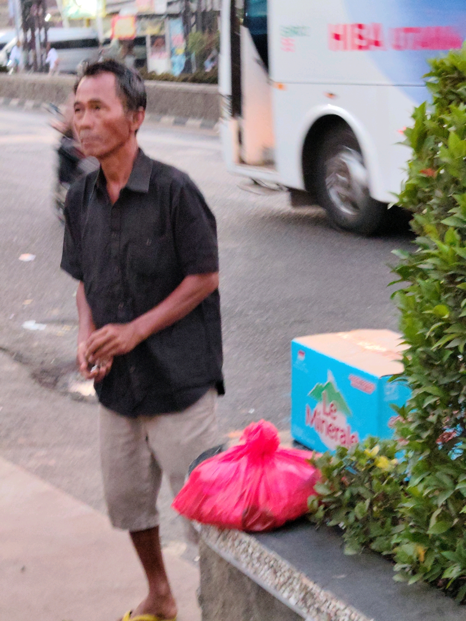 An outdoor, eye-level, medium shot captures a man of Southeast Asian descent standing in what appears to be an urban environment, possibly Purwakarta, Indonesia. The man, who is looking off to his left, is wearing a short-sleeved black collared shirt and light-colored knee-length shorts. His hands are clasped in front of him, and he is wearing yellow flip-flops. In the background, a white bus with red and blue lettering, possibly a public transportation vehicle, is partially visible, along with other out-of-focus figures and structures suggesting a street scene. To the right of the man, a bright pink plastic bag is situated on a concrete ledge. Next to the bag is a blue cardboard box with green lettering that appears to read "Le Minerale," a brand of bottled water commonly found in Indonesia. Dense green foliage frames the right side of the image, partially obscuring a street and sidewalk. The lighting suggests it may be late afternoon or early evening.