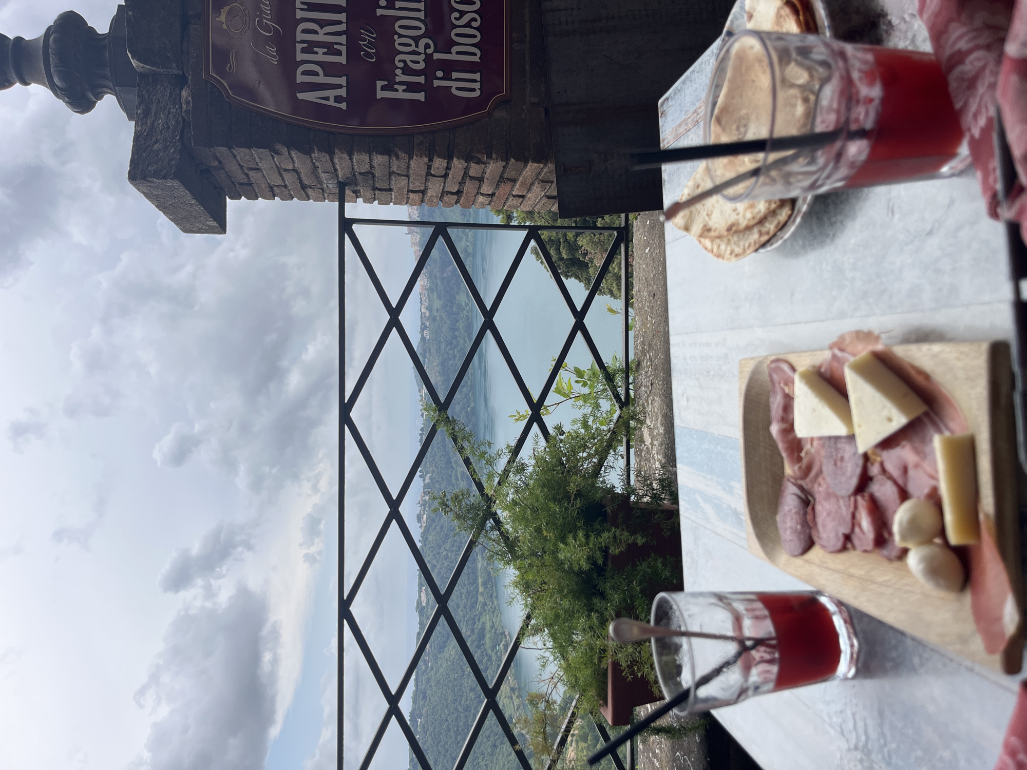 The image shows a table at an outdoor restaurant with a view of Lake Nemi in Nemi, Italy. On the table are two glasses of red juice with straws, a wooden platter with cured meats and cheese, and a plate of flatbread. In the background, there is a railing and a pot of greenery, with the lake and lush green hills beyond. A sign on a brick pillar to the right reads "la Giara Aperitivo con Fragolini di bosco". The sky is overcast with scattered clouds.
