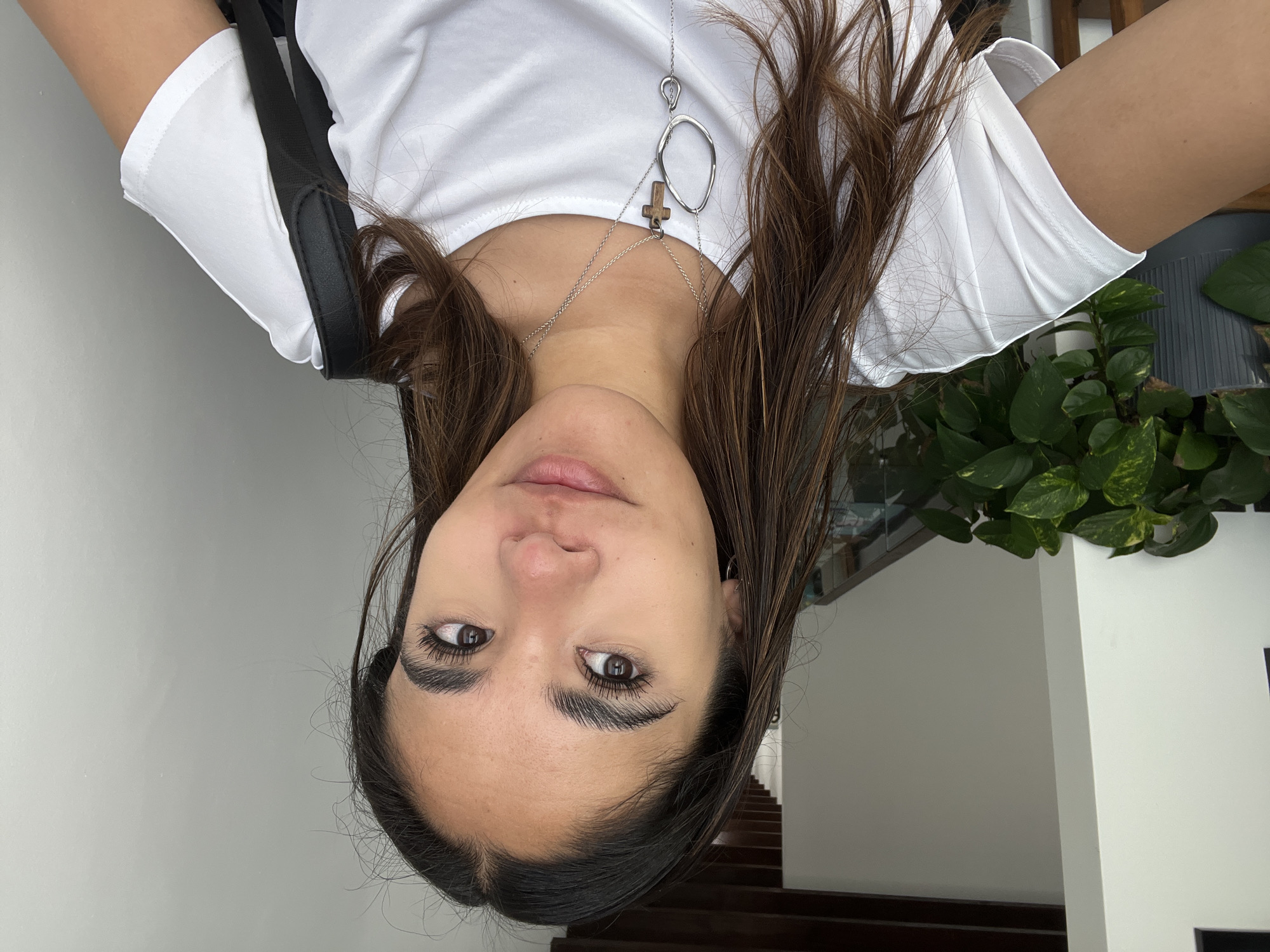 A woman with brown hair and brown eyes is posing for a selfie indoors. She is wearing a white t-shirt and a silver necklace with a cross pendant. A black bag strap is visible on her left shoulder. Behind her are white walls, a staircase with dark wood steps, and a potted plant with green leaves. The location appears to be inside a home or building in San Bernardino Tlaxcalancingo, Mexico.