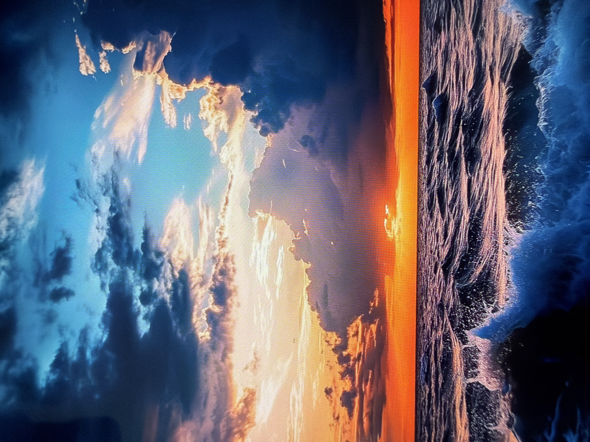 The image displays a dramatic and vibrant natural landscape, likely a sunset or sunrise over a large body of water, captured as a photograph of a digital screen.
The scene is divided horizontally by a prominent horizon line. The upper half features a dynamic sky filled with clouds. Near the horizon and towards the right, the clouds are brightly illuminated with intense hues of orange, yellow, and soft pink, indicating the presence of the low-lying sun. Further into the sky and towards the left, the clouds become denser and darker, showing cool tones of deep blue, dark gray, and purple.
The lower half of the image depicts a body of water, resembling an ocean or large lake, with noticeable wave formations. The water surface displays reflections of the warm sky, particularly near the horizon where patches of orange and pink light are visible. Toward the left, the water appears darker, with shades of deep blue and black, interspersed with the white foam and spray of breaking waves.
The sun is visible as an intense, bright glow positioned near the center-right of the horizon, casting a radiant path across the water. The overall impression is one of awe-inspiring natural beauty and a majestic display of light and color. The grid-like pixel pattern visible in the crops confirms that this is a photograph taken of a screen. There is no visible text or specific geographical cues within the depicted landscape itself.