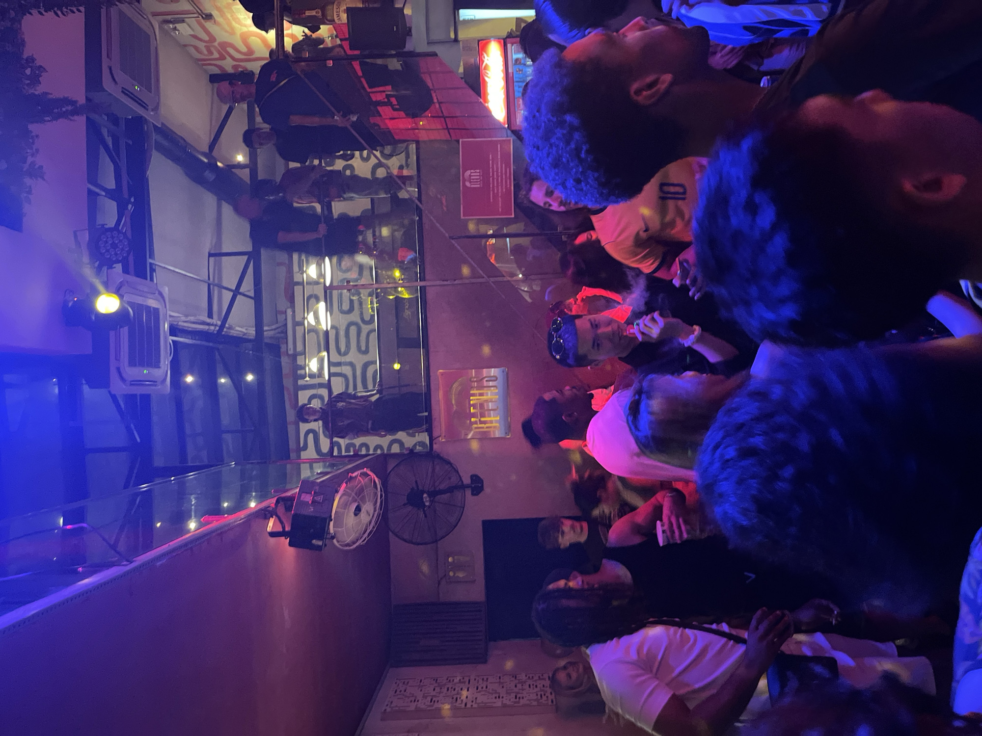 This image depicts a crowded, multi-level indoor nightclub or bar environment identified as Blends Club, located at 47 Đường Trần Phú, Hải Châu 1, Hải Châu, Đà Nẵng, Vietnam. The scene is illuminated by dominant purple and blue ambient lighting, complemented by yellow spotlights and various disco lights mounted on the ceiling and structural beams.
Numerous individuals of varying ages and apparent ethnicities are dispersed throughout the venue, standing and interacting. A significant number of patrons occupy the main floor, while others are visible on an upper balcony or mezzanine level, accessible via a staircase on the right. People are observed holding drinks in cups, and one individual in the lower-left foreground is actively vaping, exhaling a plume of vapor. Another person in the mid-ground wears an orange sports jersey with the number "10" on the back.
The club features an industrial aesthetic with exposed black metal beams and ductwork. Several large, black wall-mounted fans are visible, along with multiple air conditioning units on the ceiling. A prominent red sign on the mid-right wall displays the "BLENDS" logo and additional, smaller text. Below this sign is a red Coca-Cola cooler. A bar area is partially visible on the upper right, where bottles, including a Jameson whiskey bottle, are present. Abstract patterns or graffiti adorn some wall sections, and artificial greenery hangs from the ceiling in certain areas. The overall atmosphere appears lively and social.