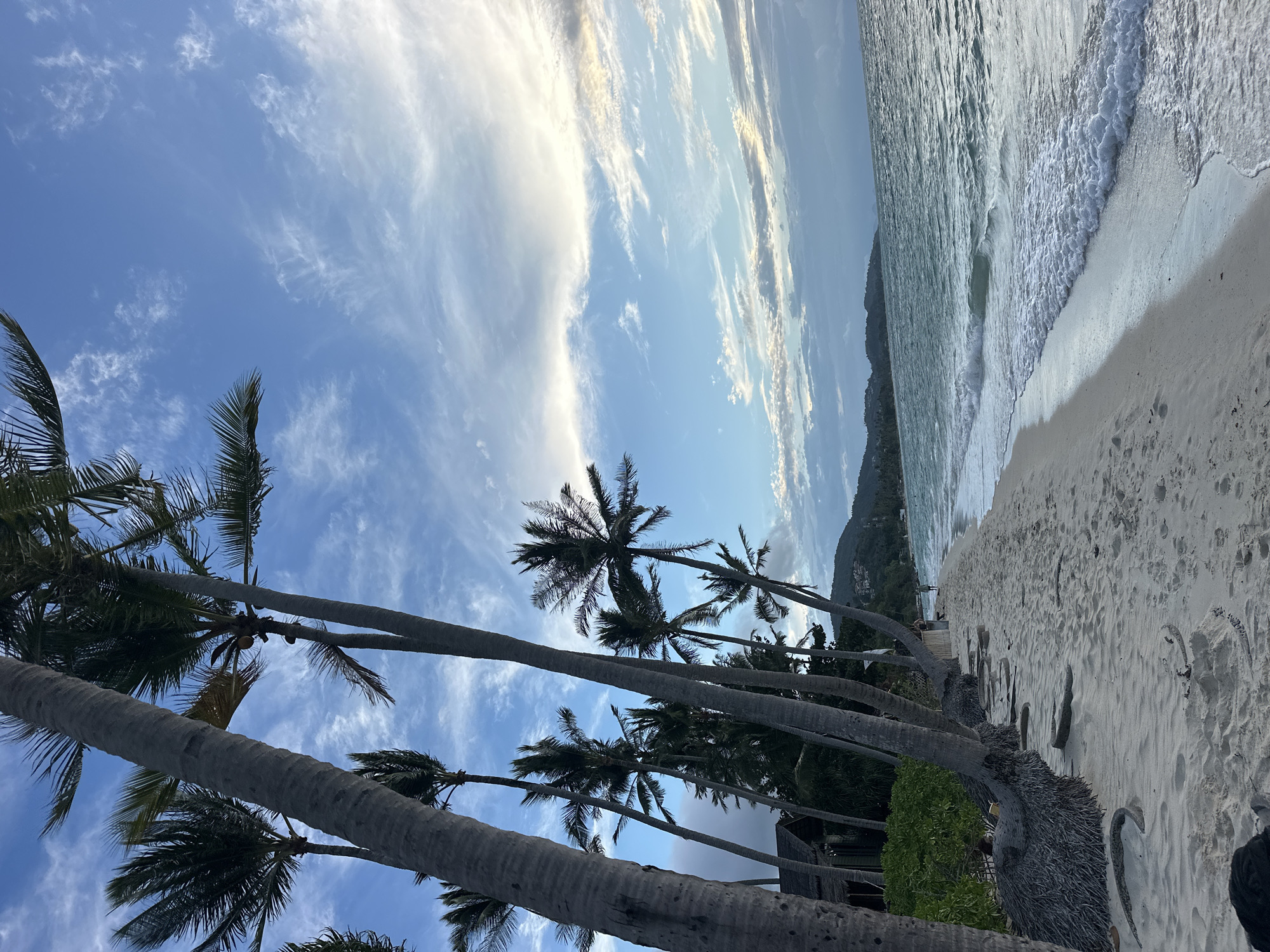 The image captures a tropical beach scene in the Ko Samui District of Thailand. A row of tall palm trees with textured trunks lines the left side of a sandy beach. Their fronds reach towards a blue sky filled with wispy white clouds. In the background, the turquoise ocean waves gently lap at the shore, creating a foamy white edge. Beyond the water, a distant, verdant hillside with scattered buildings can be seen. A small, thatched-roof structure is partially visible nestled among the trees on the left. Two people are walking on the beach in the mid-distance, and some scattered driftwood and debris are present on the sand. The overall lighting suggests it might be late afternoon or early morning.