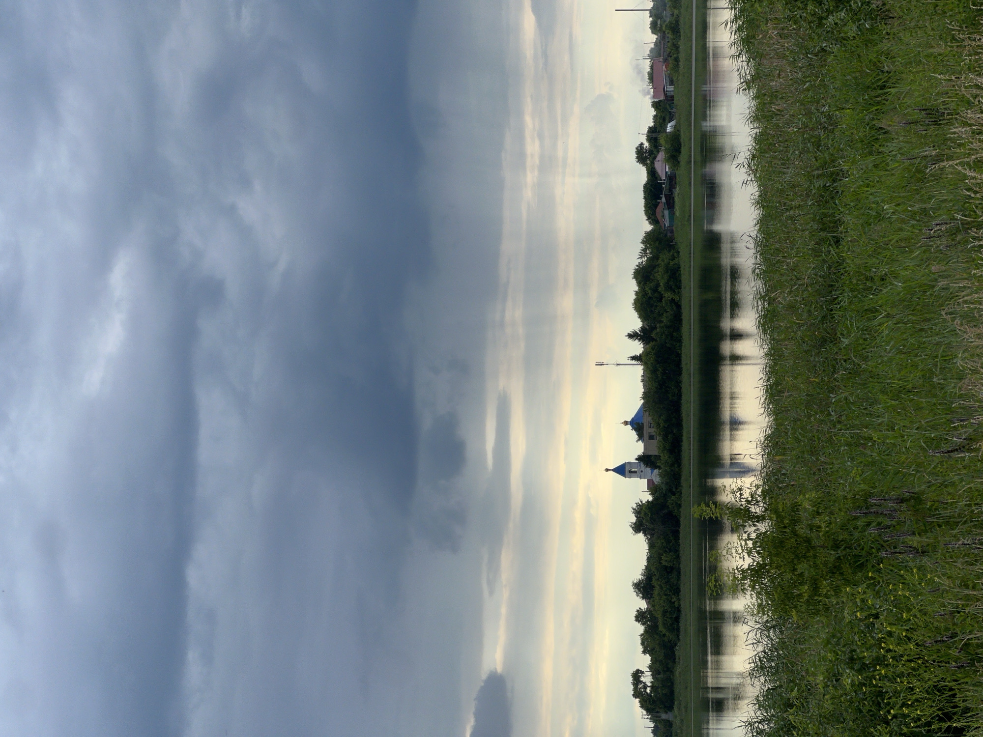 The image depicts a tranquil natural scene of a wide river and its verdant banks under a dramatic, overcast sky. In the background, across the river, a small settlement is visible, characterized by dense green trees, a few houses, and two distinct church buildings. One church features a tall, slender white belfry topped with a blue dome and a golden cross, while the other is partially obscured but also shows a blue dome. The sky is filled with heavy, dark gray clouds, with lighter patches of sky and streaks of sunlight breaking through in the distance, creating a diffused, atmospheric light. The river itself is calm, reflecting the sky and the distant shoreline with a muted sheen. In the foreground, the riverbank is densely covered with tall green grass and some wildflowers, creating a natural, untamed border to the water. The overall impression is one of peaceful, rural serenity, possibly captured during or just before a rainfall, given the cloud formations. The location is identified as Pivkino, Russia.