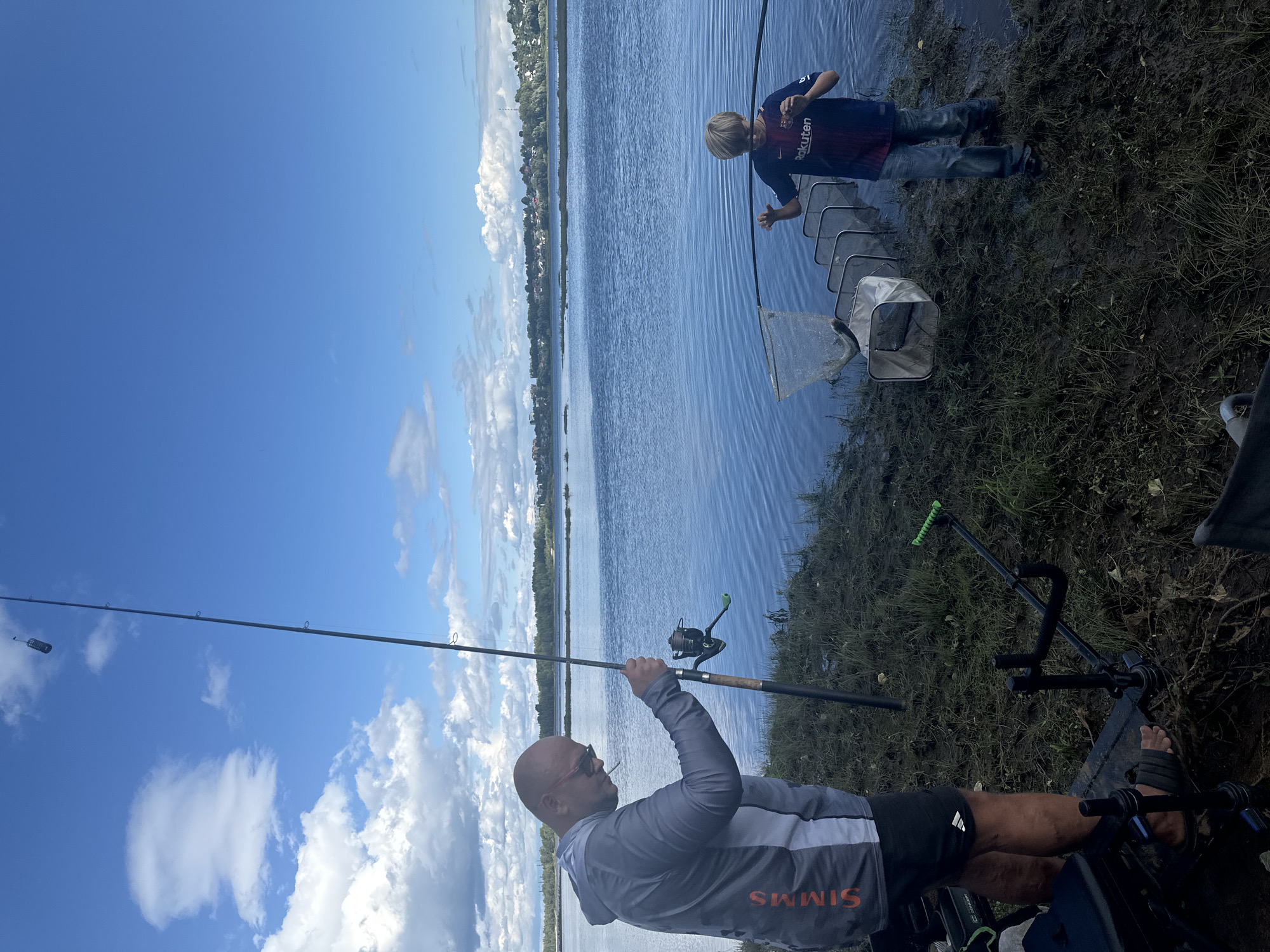 An adult male with sunglasses and a gray and white fishing shirt is standing on the bank of a body of water, holding a fishing rod. To his right, a young male in a blue and maroon soccer jersey is holding a fishing net with a fish in it, and has a series of connected cages in the water in front of him. The background shows a wide expanse of water with a treeline and buildings on the opposite shore. The sky is blue with white clouds. The foreground shows grassy and muddy terrain with fishing equipment.