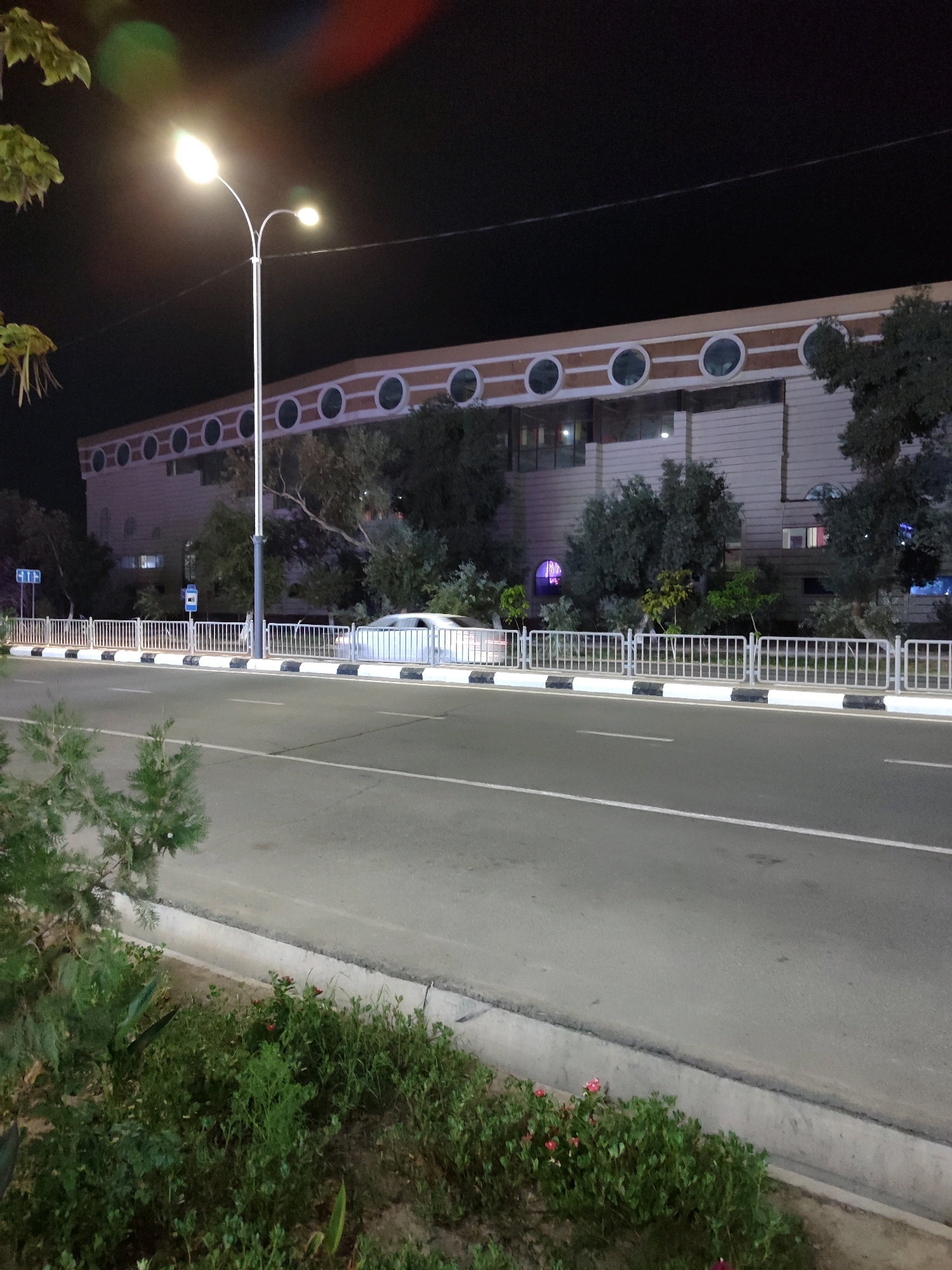 The image shows a nighttime street scene in Nukus, Uzbekistan, likely near a public building or complex. A prominent street lamp with two lights illuminates the foreground. A paved road with white lane markings stretches into the distance. A white metal fence with black and white bollards separates the road from a sidewalk. Beyond the fence, trees and bushes are visible, and behind them, a large building with a facade featuring a series of circular windows can be seen. A white car is parked near the building. The sky is dark, indicating it is nighttime.