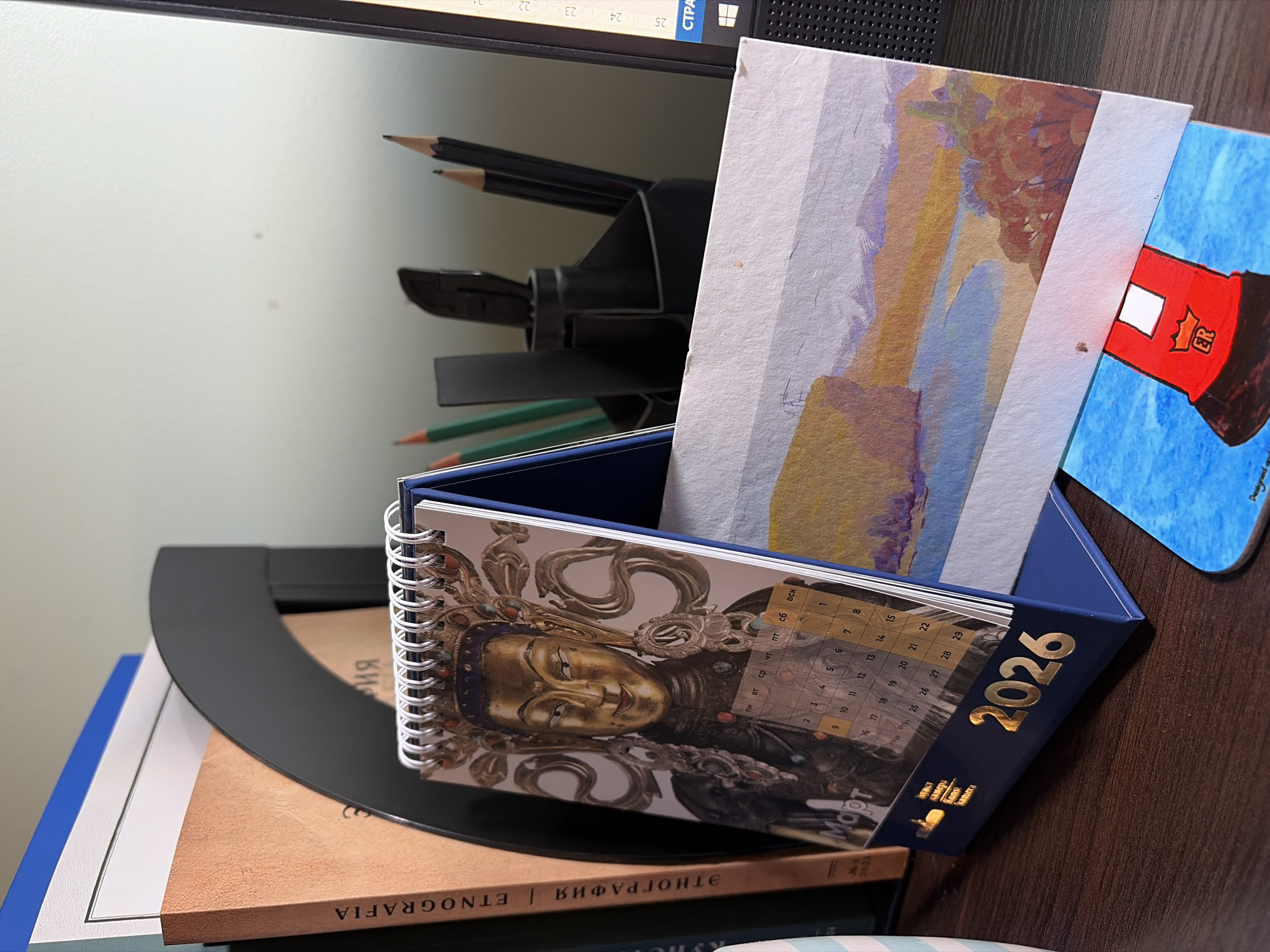 The image depicts a desk setting. A 2026 desk calendar is prominently featured with an image of a golden Eastern religious figure on its cover, displaying a calendar grid for March. The calendar is navy blue with the year "2026" printed in gold. Behind the calendar are various postcards or artworks and writing utensils. Underneath the calendar are books, one titled "ETNOGRAFIA," potentially referring to ethnography. The setting appears to be indoors, likely an office or study. The presence of Cyrillic text hints at a location in a Slavic-speaking region, likely Russia or a former Soviet republic. There is no indication of the location in the city of Saint Petersburg, Russia.