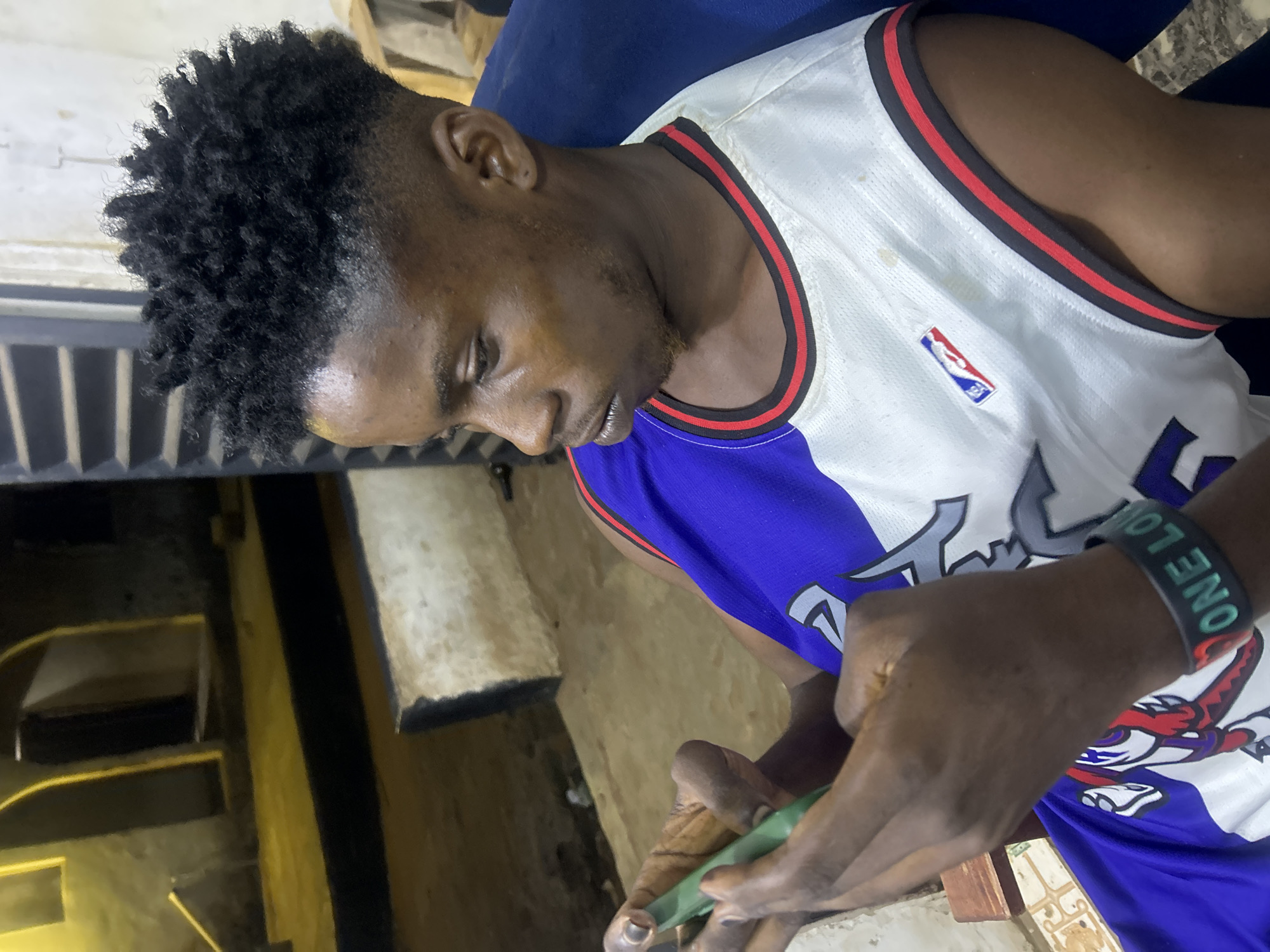 A young Black man is depicted in a medium shot, seen from the chest up, looking downwards and to his right, deeply engrossed in an activity. He has short, dark, curly hair with faded sides and a neat, short beard or goatee. He is wearing a white basketball jersey with red and black trim around the neck and armholes. The jersey features the NBA logo on the front and a blue section at the bottom, suggesting a Toronto Raptors design with a partial raptor logo visible.
In his hands, he holds a small, rectangular, green object, possibly a smartphone or a similar electronic device, which he appears to be operating with his thumbs. On his left wrist, he wears a black wristband with the words "ONE LOVE" inscribed in green text.
The setting appears to be an indoor or semi-enclosed space, characterized by muted, earthy tones. The background shows textured walls or structures in shades of yellow and brown, with darker, shadowed areas suggesting an interior environment with ambient lighting. The man's expression conveys focus and concentration on the object in his hands. The location context provided is Ikorodu, Nigeria.