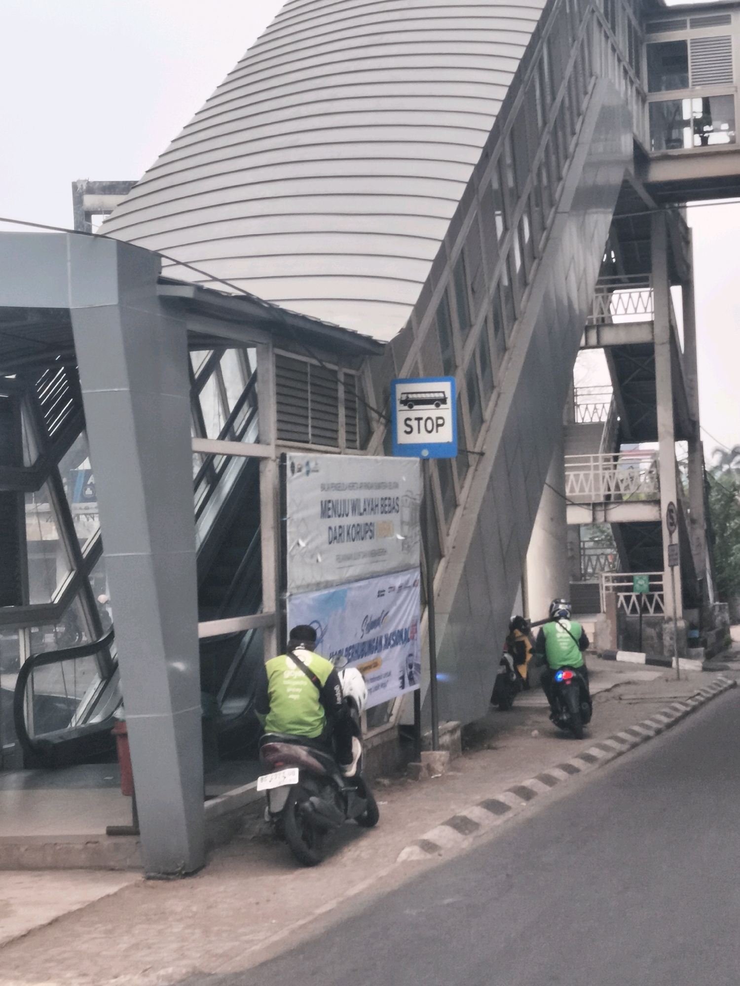 The image depicts an outdoor scene in Palembang, Indonesia, featuring a modern pedestrian bridge and an escalator entrance. In the foreground, a person wearing a bright green vest, likely a delivery driver, is seated on a motorcycle. Another motorcycle is partially visible to the left, and a third is parked further down the street. Two banners are displayed on a structure next to the escalator. One banner features a bus stop sign, indicating a transportation hub. The architecture is contemporary, with a large, curved, ribbed structure forming the main element of the pedestrian bridge, contrasted with metal scaffolding and walkways. The road in front of the pedestrian bridge is paved, with a striped curb. The sky is overcast, suggesting a cloudy day. The overall impression is of a functional urban environment with public transportation infrastructure.