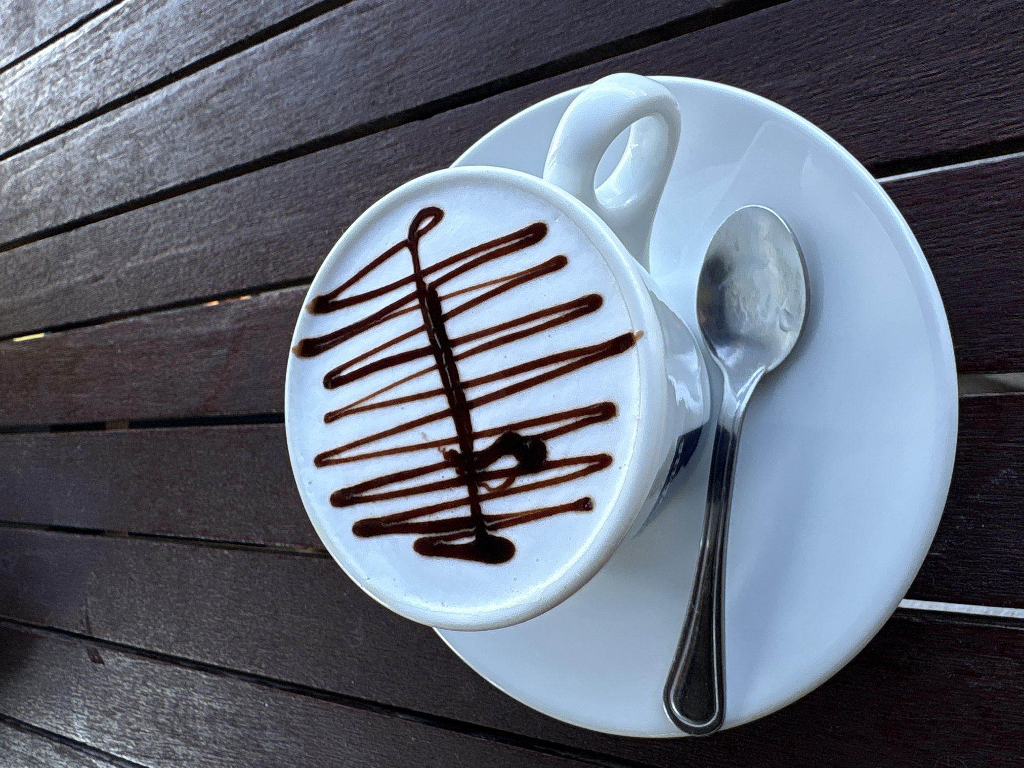 A top-down shot presents a cup of coffee placed on a white saucer with a spoon. The coffee features a white frothy surface decorated with a brown chocolate syrup design. The cup itself is white with a curved handle. The spoon rests on the saucer alongside the cup. The composition is set on a dark, horizontal wooden surface with visible lines and grain. The location is in the city of Bar, Montenegro.