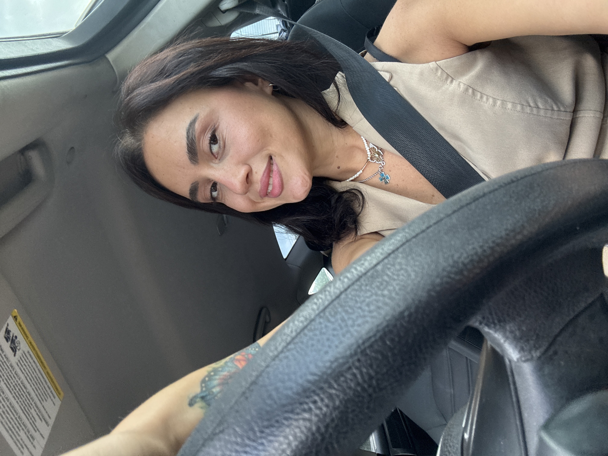 A woman is sitting in the driver's seat of a car, holding the steering wheel. She is looking towards the camera and smiling. She has dark hair and is wearing a light-colored vest and a necklace. A seatbelt is visible across her chest. The interior of the car is visible, including the dashboard and the passenger seat. The scene appears to be in an urban environment, but no specific landmarks are visible to identify the location as Tbilisi, Georgia.