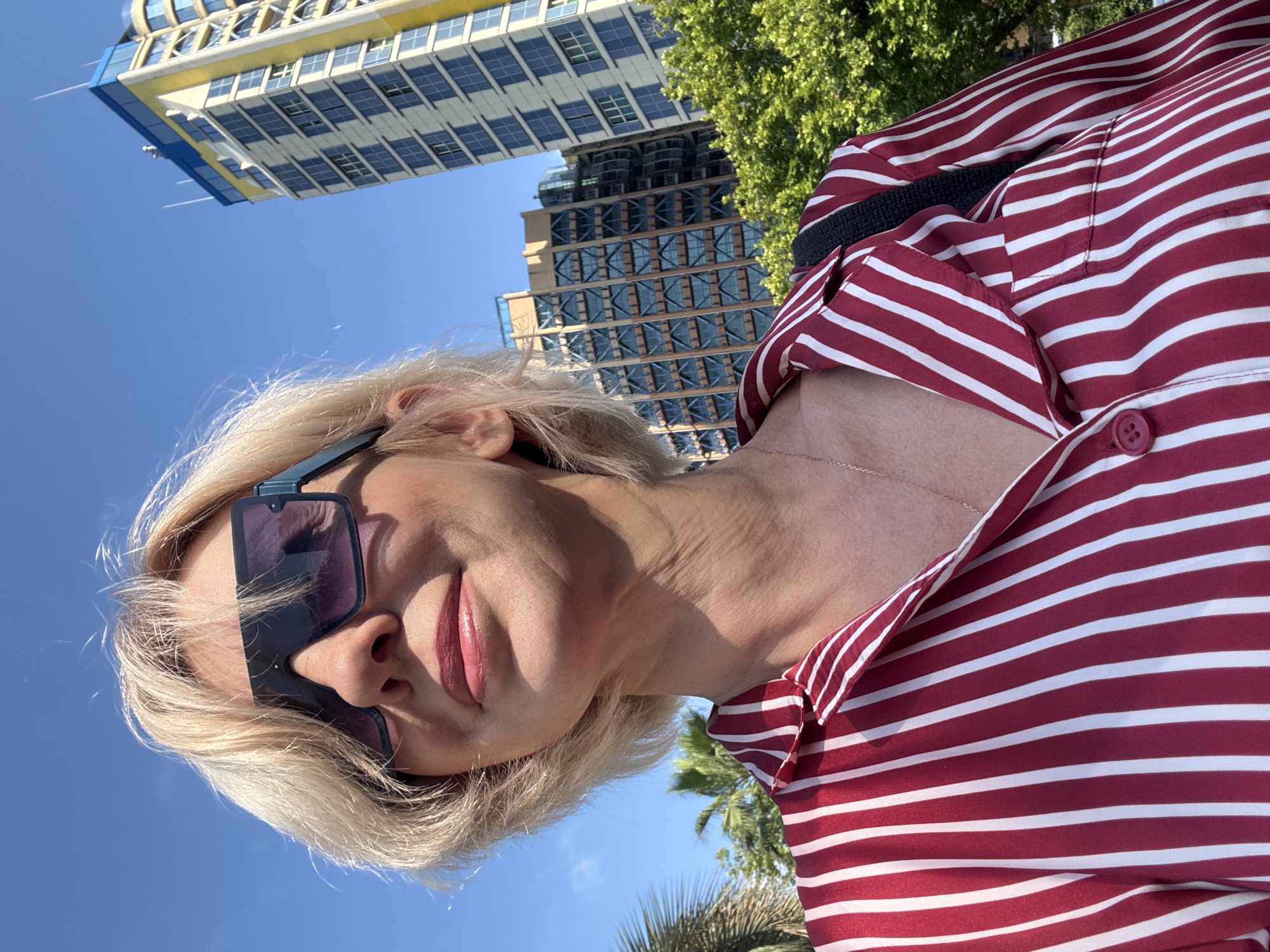 A young woman with blonde hair and sunglasses is captured in a close-up, low-angle shot. She wears a red and white striped shirt and a delicate necklace. Her expression is serene and she is looking directly at the camera.
Behind her, two modern buildings with glass facades are visible, suggesting an urban setting. Green trees and a clear blue sky frame the scene, indicating it is daytime. The sunlight suggests it's a warm day. The overall mood is relaxed and sunny.
The media file appears to be a selfie taken outdoors. The context provided places the location as Sochi, Russia. No visible text is present in the image.