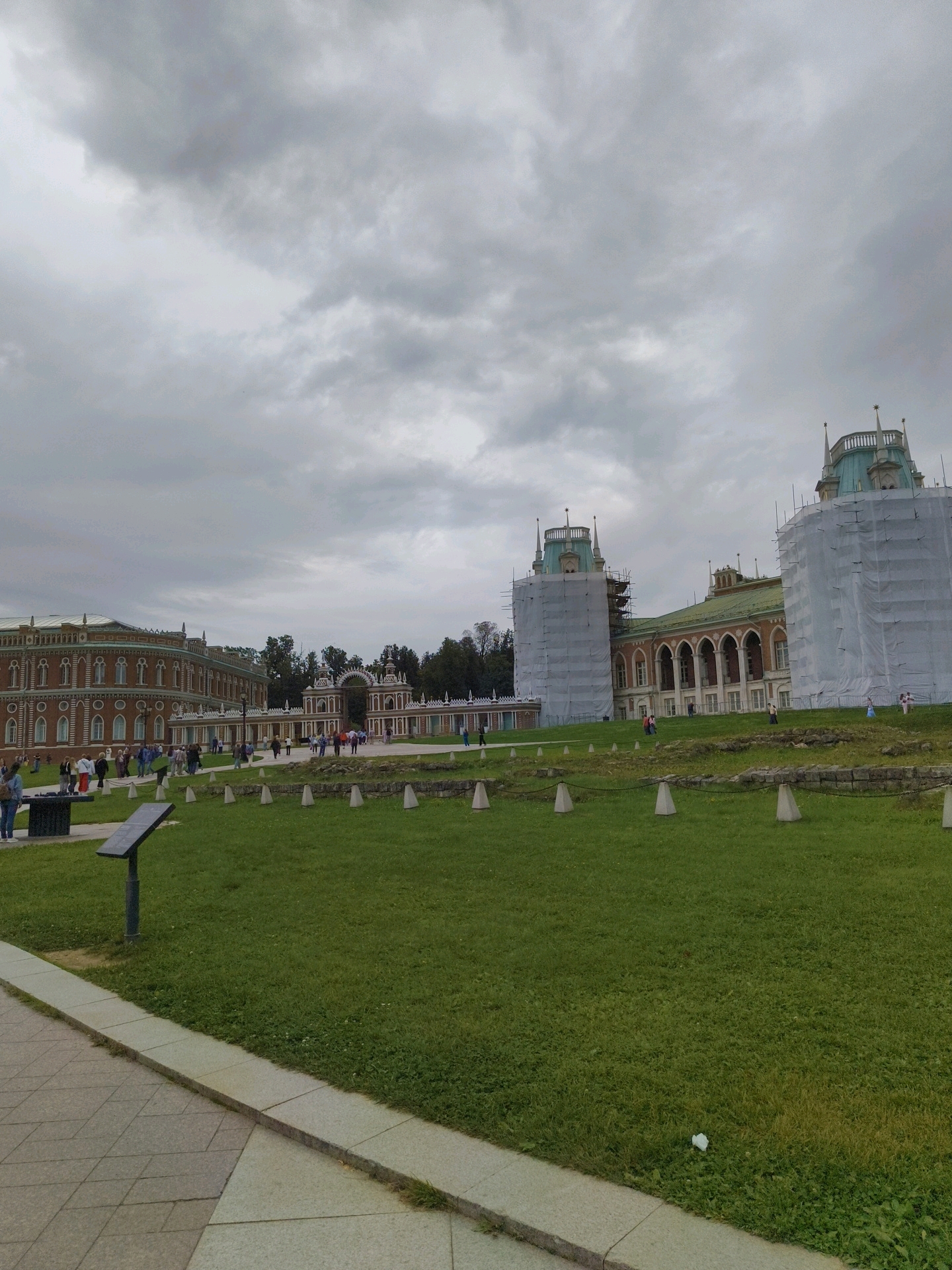 The image depicts a wide view of the Tsaritsyno Palace complex in Moscow, Russia, on an overcast day. The palace buildings are visible in the background, with one section covered in white scaffolding and sheeting, suggesting ongoing construction or renovation. In the foreground, a large expanse of neatly mown green grass occupies most of the frame, with a paved walkway on the left edge. Several small, light-colored conical objects are arranged in a line across the lawn, spaced evenly apart. A dark information kiosk stands on the left side of the lawn. In the middle ground, a row of people are seen walking along a path in front of the palace structures, which feature ornate architectural details. The sky is filled with heavy, grey clouds, contributing to a muted and somber atmosphere.