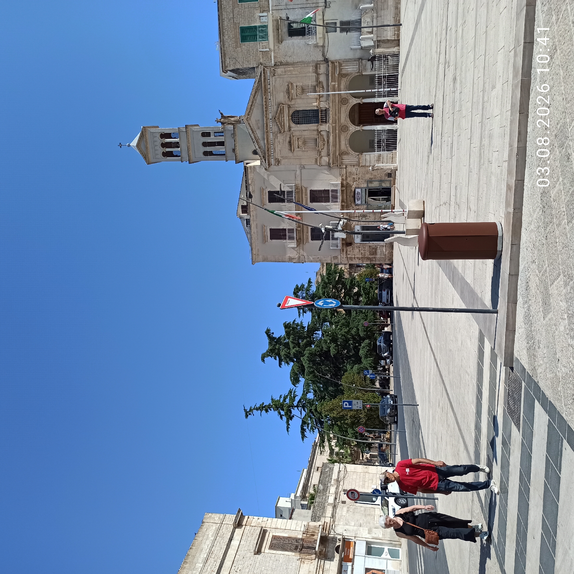 The image shows a street scene in Ruvo di Puglia, Italy, on a sunny day. In the foreground, two people are walking across a pedestrian crossing. A woman in a black dress and a man in a red shirt and jeans are walking side by side. Behind them, a brown trash receptacle stands on the paved square. In the background, a large building with a bell tower, likely a church, dominates the skyline. The building has classical architectural features and a statue of Christ at its apex. Several cars are parked or driving on the street. Trees line the area, and Italian flags are visible on flagpoles. The sky is clear blue. A timestamp "03.08.2026 10:41" is superimposed on the lower right corner of the image.