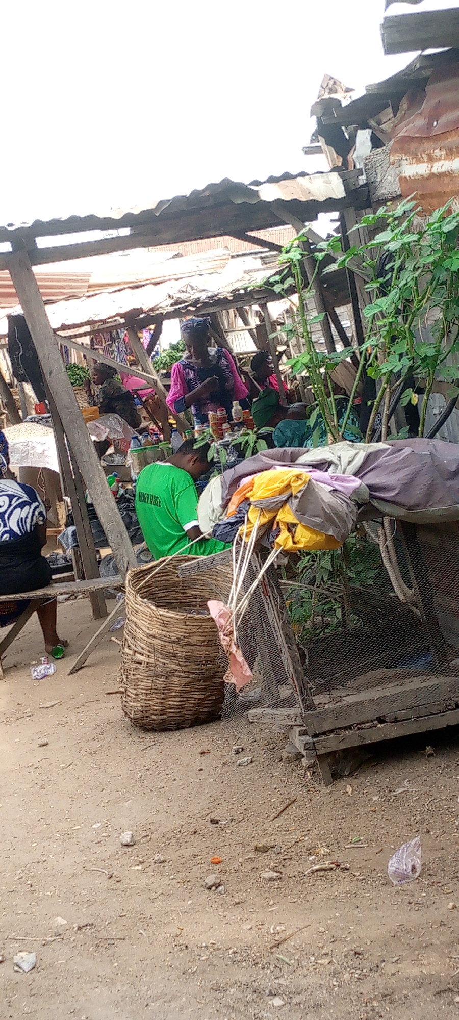 The image depicts a marketplace in Zango-ndaji, Nigeria. Several individuals are present, including a person in a green shirt with the word "PENTECOST" visible on the back. Women in traditional African clothing are seated at stalls, seemingly selling goods. Objects in the scene include a large woven basket, a chicken coop with clothes draped over it, and various market items on display at the stalls. The environment is characterized by rudimentary wooden structures and a dirt ground.