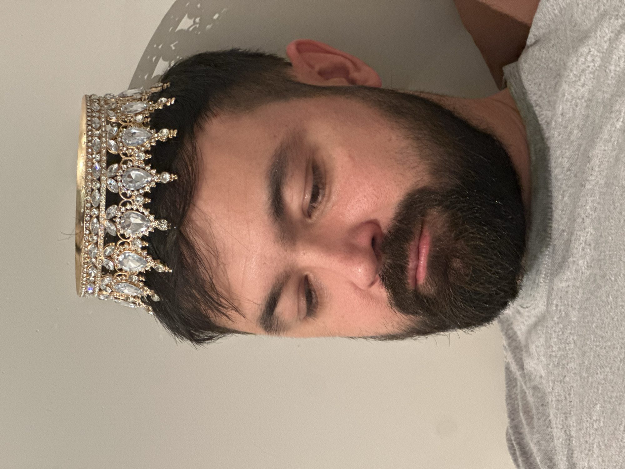 A close-up, portrait-oriented image shows a man lying down, viewed from a slightly overhead perspective. He has dark hair and a full beard and mustache. His eyes are closed, giving him a calm and relaxed expression. On his head, he wears a prominent, gold-toned crown richly adorned with numerous clear, sparkling gemstones, primarily teardrop and round shapes. A light grey t-shirt is visible on his left side. The background consists of a plain, light-colored wall, indicative of an indoor setting. Subtle shadows are cast on the wall above his head, suggesting an artificial light source. No text is visible in the image. The scene captures a quiet, introspective moment, possibly one of rest or contemplation, taking place in Khimki, Russia.