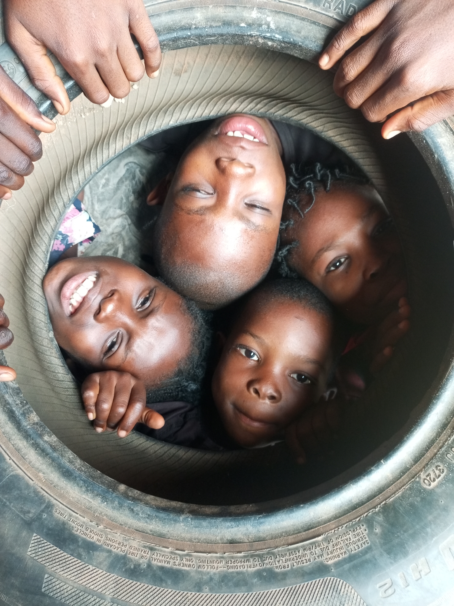 A high-angle, close-up shot captures four Black children crammed inside a large, dirty tire, which appears to be a spare tire from a truck or heavy-duty vehicle, at a location in Gombe, Nigeria. The children's faces are pressed together, looking upwards towards the camera. One child, in the upper left quadrant of the image, has their eyes closed and mouth open. Another child, positioned below and slightly to the left of the first, has their eyes wide open, smiling, and displaying their teeth. To the right of this child is another child with their eyes open, looking towards the camera. The fourth child is located in the upper right quadrant, with their eyes open and looking slightly off-camera. The children are wearing casual clothing. The tire itself is worn and shows signs of dirt and dust. The interior of the tire is dark, except for where the children's faces are illuminated by the camera's flash. The exterior of the tire, visible around the edges of the frame, displays tire markings and tread.