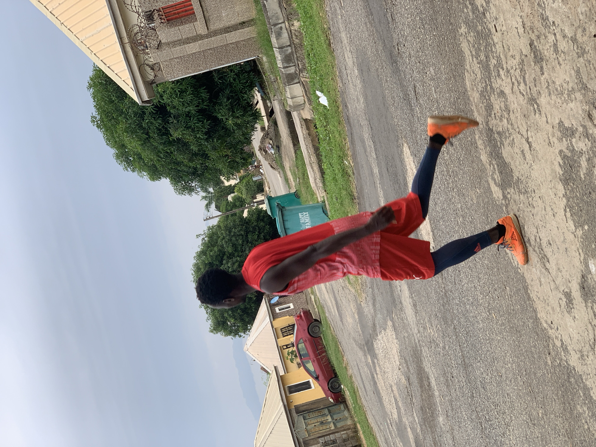 In Abuja, Nigeria, a young man with dark skin and black hair, wearing a red sleeveless shirt with white stripes and red shorts over dark blue leggings, is captured mid-stride. His right leg is extended forward, and his left leg is bent at the knee, with orange athletic shoes on both feet. He is jogging on a paved road with some patches of wear and tear.
The environment appears to be a residential area. To the left of the man, there are two houses. The closer house is painted yellow with a white roof and a metal gate. Further back, a red sedan is parked in front of a house with a white roof. Behind the houses, a dense green tree canopy dominates the middle ground. To the right of the man, a beige building with a yellow roof and a stone facade is visible, with barbed wire along the top of its wall. In front of this building, there is a blue garbage bin with white lettering. A concrete drainage channel runs along the side of the road, bordered by grass. The sky is overcast with a light grey hue. In the distance, mountains can be seen under the hazy sky.