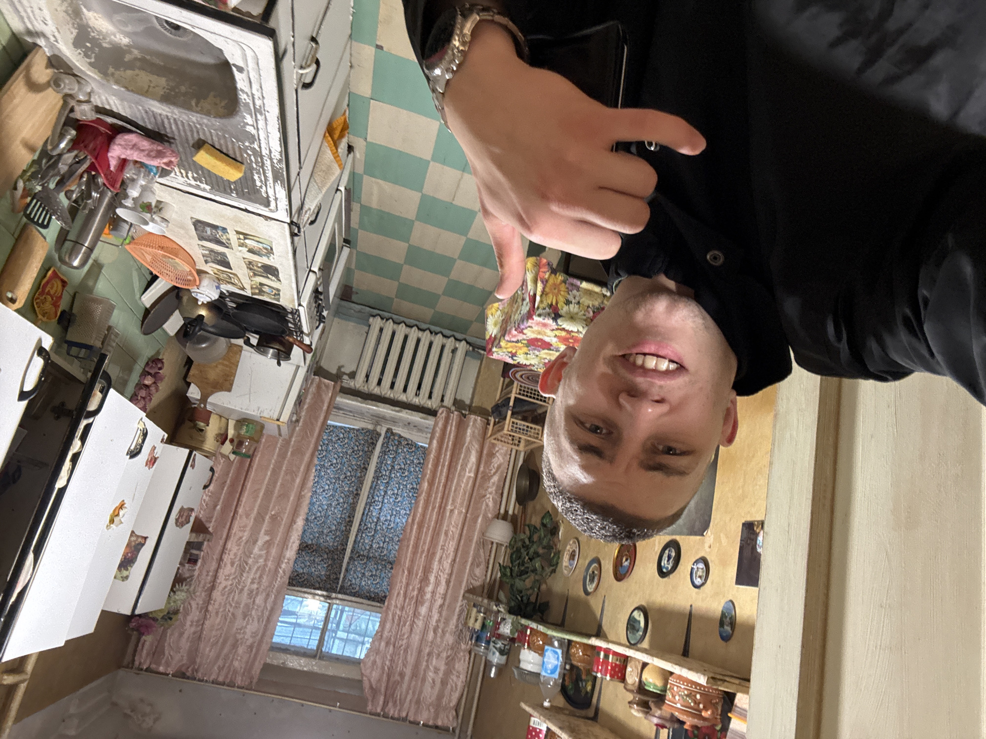 A man is taking a selfie in a kitchen. The kitchen features a white sink, a white stove, and a checkered floor. A radiator is visible near a window with pink curtains. The walls are decorated with small plates. The location is Unknown, Unknown.